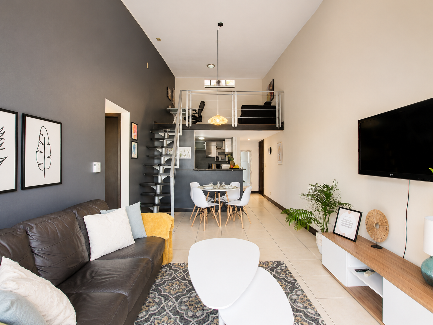 Open-concept living room and dining area with a high ceiling, dark and light wall colors, a brown leather sofa with pillows, a white modern chair, a patterned rug, a small round dining table with four white chairs, a floating staircase, an upper loft