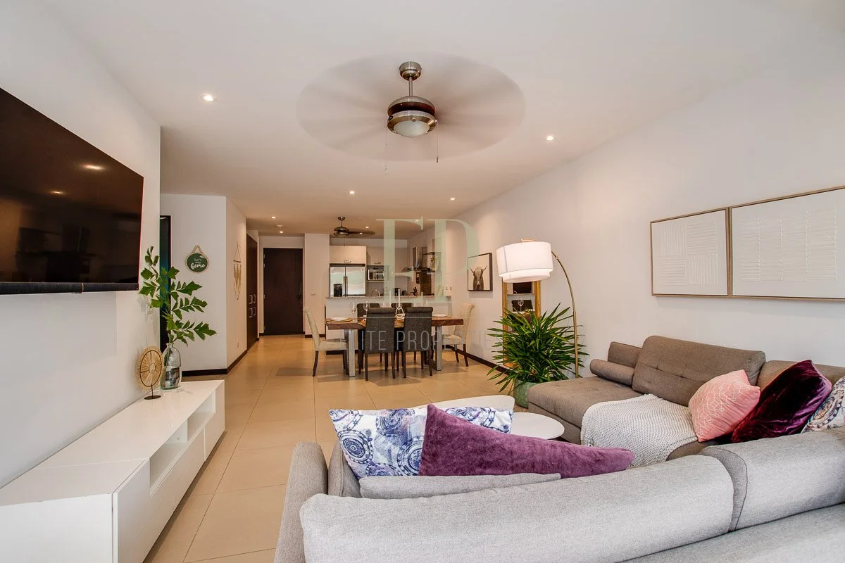 Living room with gray sectional sofa, colorful pillows, floor lamp, wall art, and a large flat-screen TV on the wall. Open dining area and kitchen in the background, with a dining table and chairs.