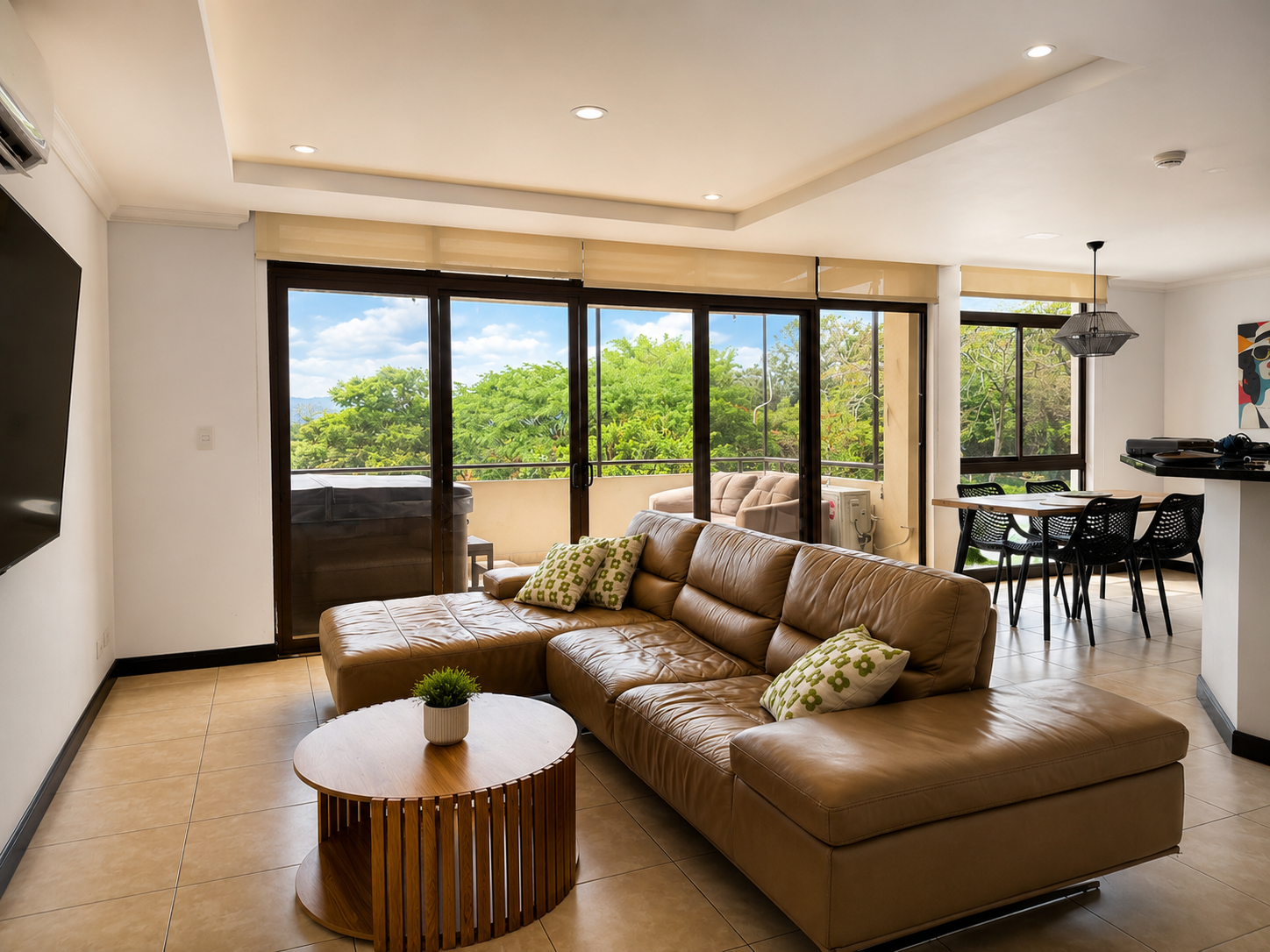 Living room with a large brown leather sectional sofa, green patterned pillows, a small round wooden coffee table with a potted plant, a wall-mounted TV on the left, and sliding glass doors leading to a balcony with outdoor furniture and lush greener