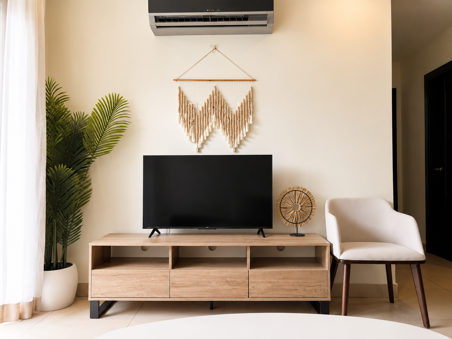 Living room with a flat-screen TV on a wooden stand, a large green plant in a white pot on the left, a decorative fan-shaped lamp on the right, a beige armchair, a wall-mounted air conditioning unit, and a wall hanging made of beads.