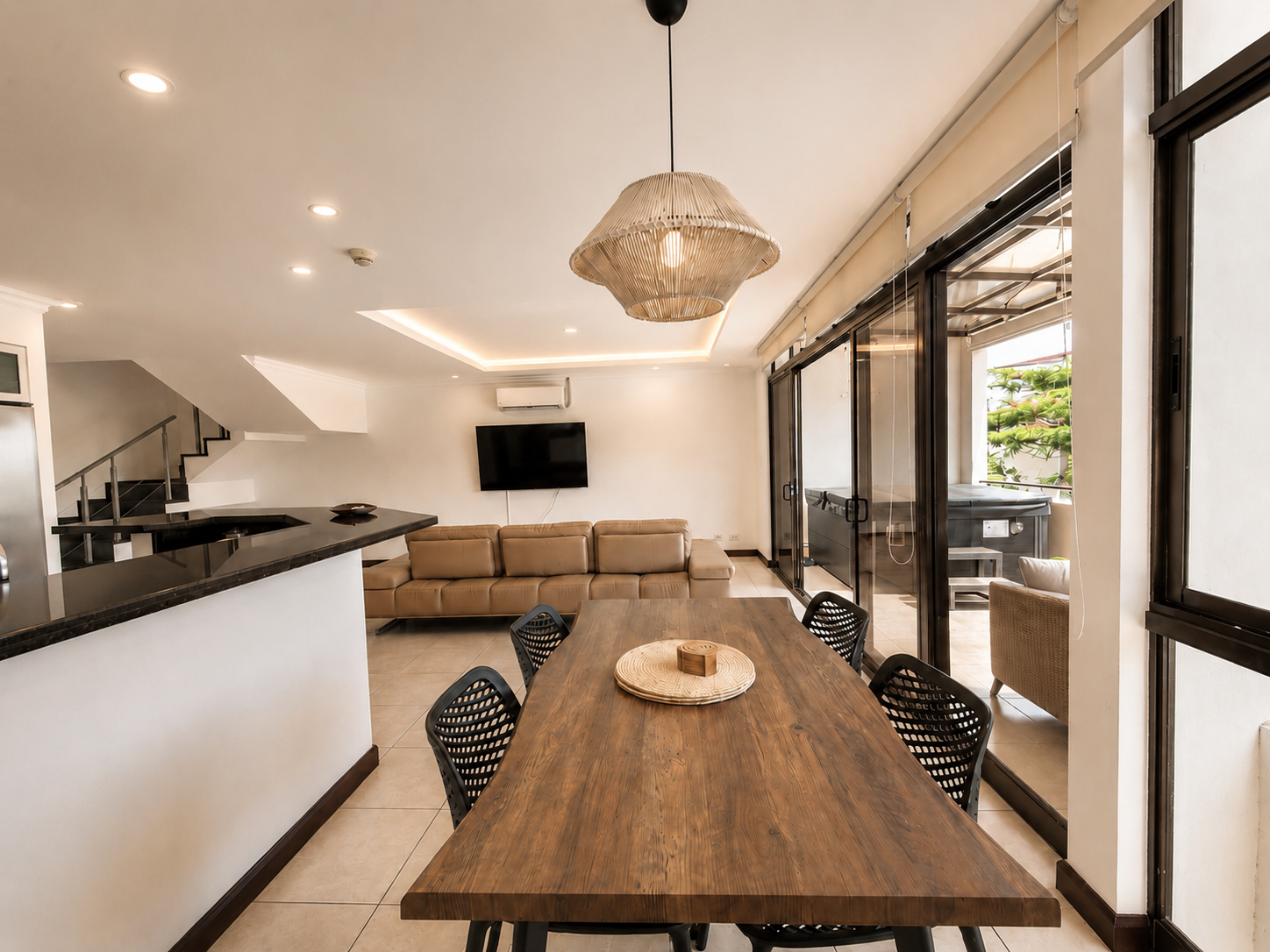 Open-concept living and dining area featuring a wooden dining table with six black chairs, glass sliding doors leading to an outdoor space, a beige sectional sofa facing a wall-mounted TV, and a decorative woven pendant light hanging from the ceiling