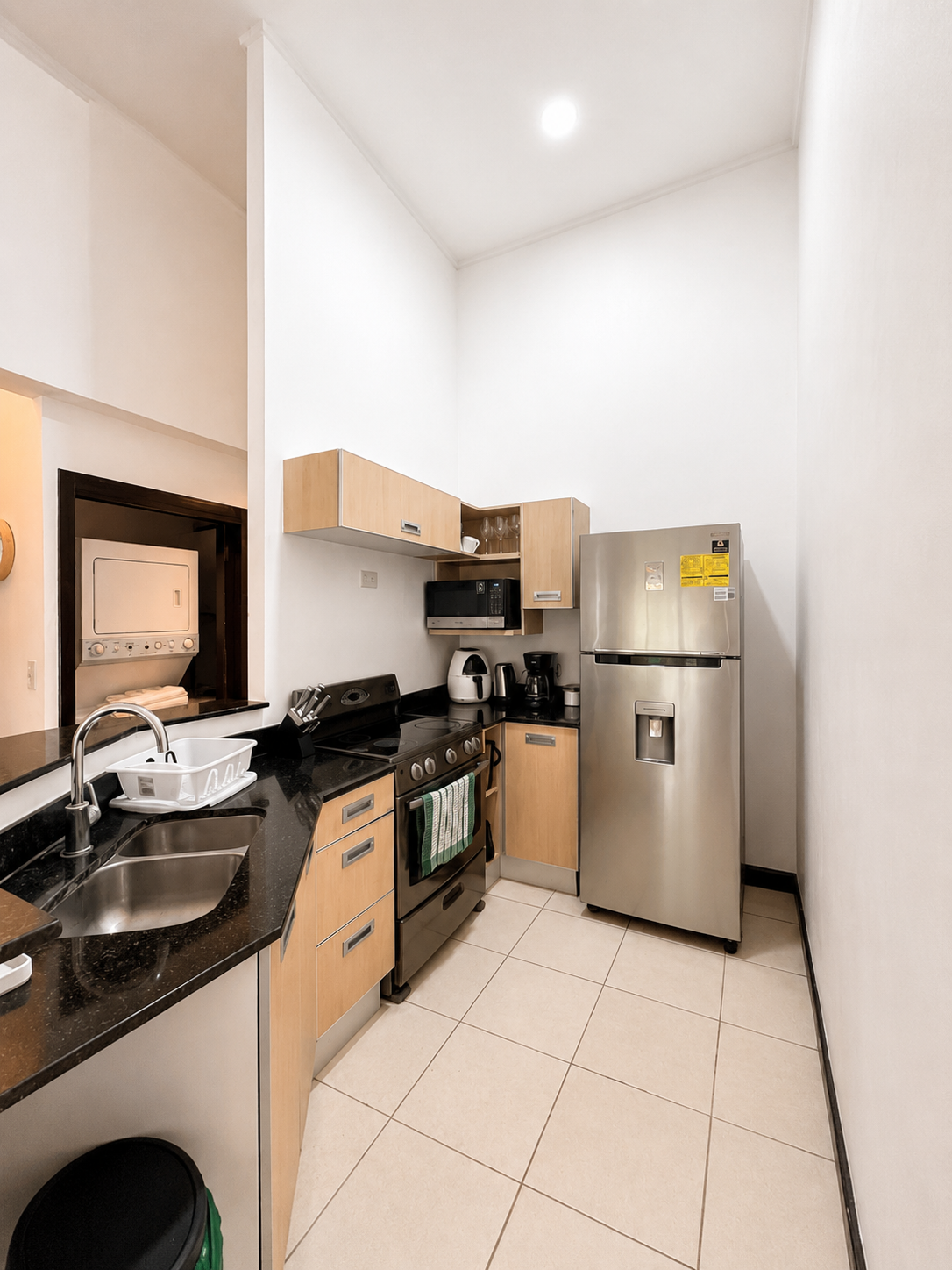 A compact kitchen with light wood cabinets, black countertop, stainless steel appliances, including a refrigerator, stove, microwave, and coffee maker, with white tiled flooring and white walls.
