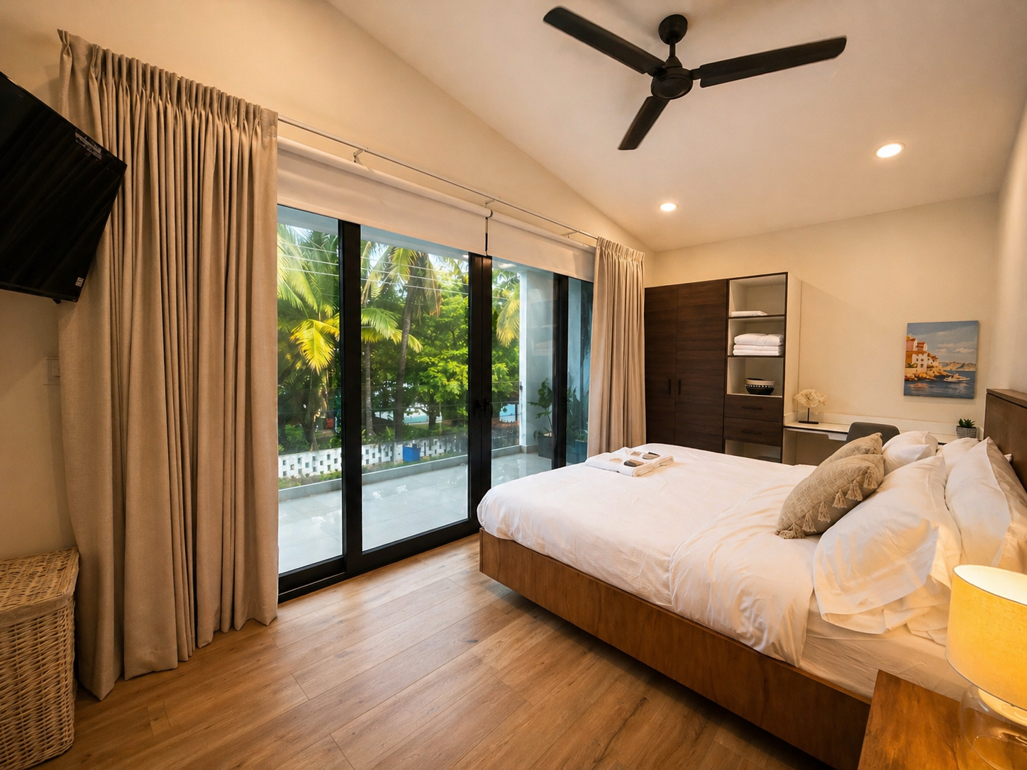 A bedroom with a large sliding glass door showing a view of lush green trees outside. The room has a bed with white bedding and pillows, a wooden side table, a dark wood wardrobe, a paint of a coastal scene on the wall, a desk with a small lamp, and 