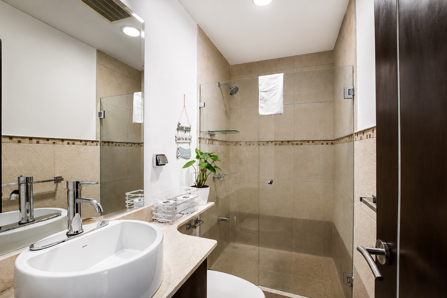 A modern bathroom featuring a white vessel sink with a chrome faucet, a large mirror, a potted plant, and a shower area with a glass partition, beige tiles, and a wall-mounted showerhead.