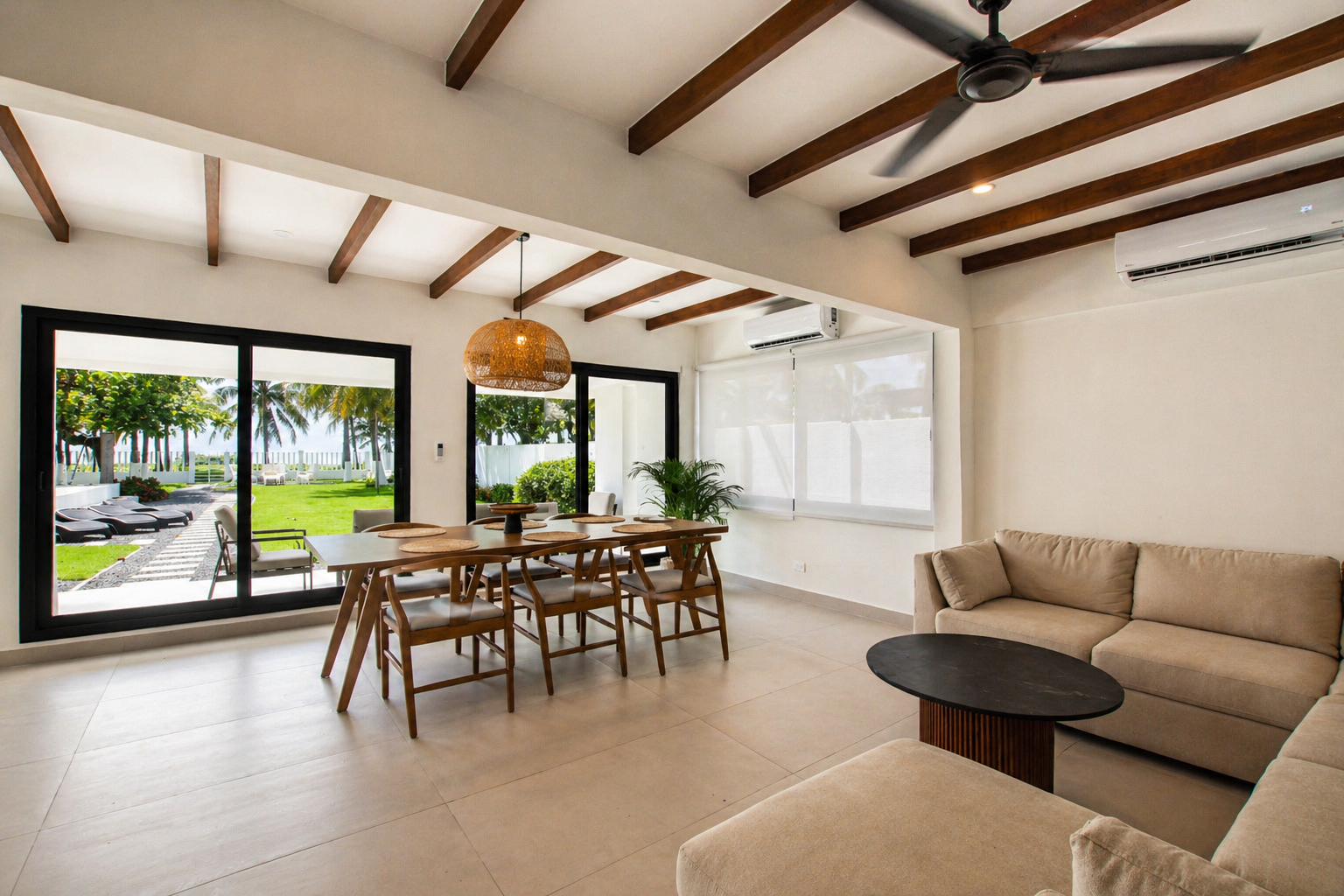 Bright living room with large sliding glass door opens to outdoor garden, wooden dining table with chairs, beige sectional sofa, black coffee table, white walls, ceiling fan, air conditioning units, and palm trees visible outside.