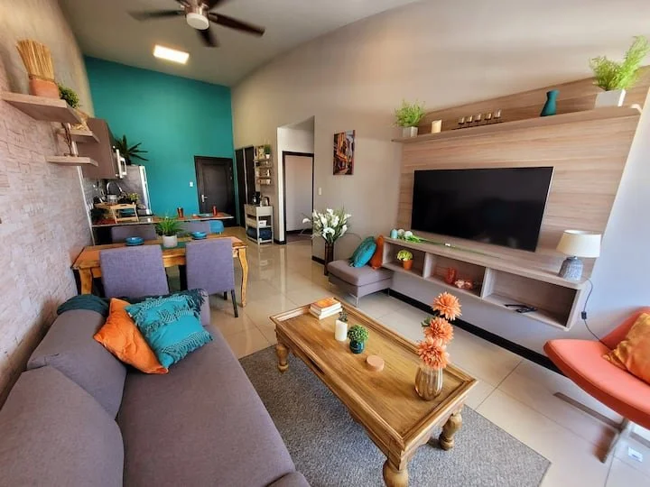 Living room with a gray sofa, colorful pillows, a wooden coffee table with flowers, a flat-screen TV, and a dining area with a wooden table and purple chairs, decorated with plants and artwork.