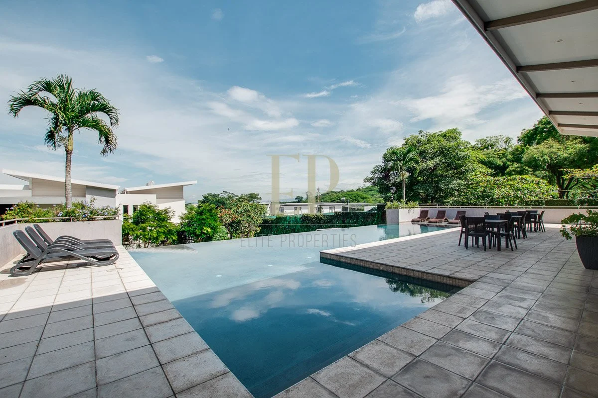 Outdoor patio with a swimming pool, lounge chairs, a table with chairs, lush greenery, and a clear blue sky.