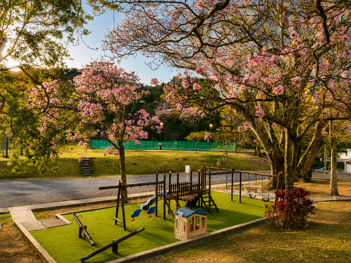 A park with pink blossom trees, a small plastic playhouse, swings, trees, and a tennis court in the background.