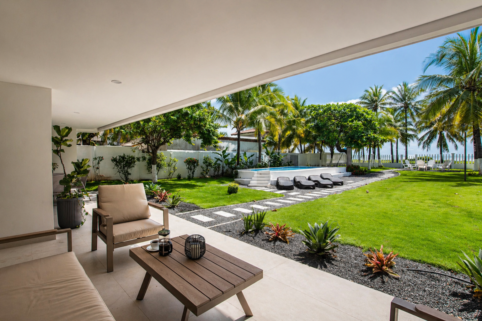 View of a tropical backyard with a pool, lounge chairs, palm trees, and a seating area on a shaded patio.