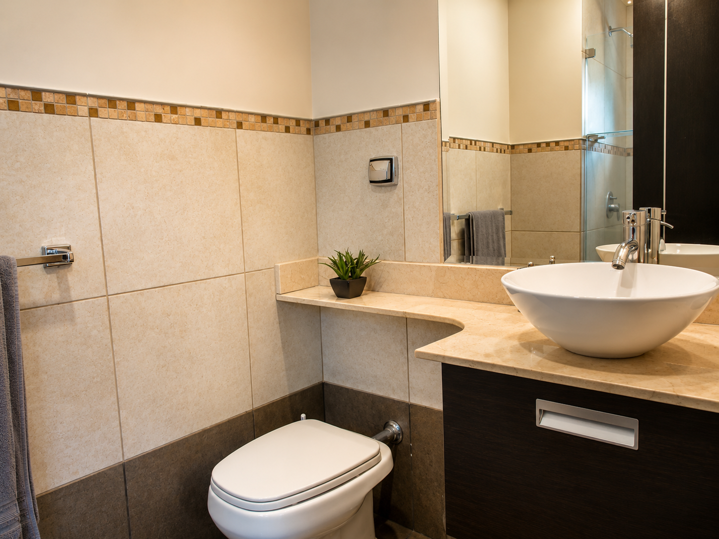A modern bathroom with a beige tiled wall, a large mirror, a white vessel sink with chrome faucet, a small potted plant on the marble countertop, and a visible shower area with glass door.