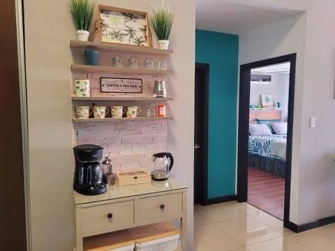 Coffee station with a coffee maker, glass jar, and teapot on a small cream-colored table against a white brick wall.