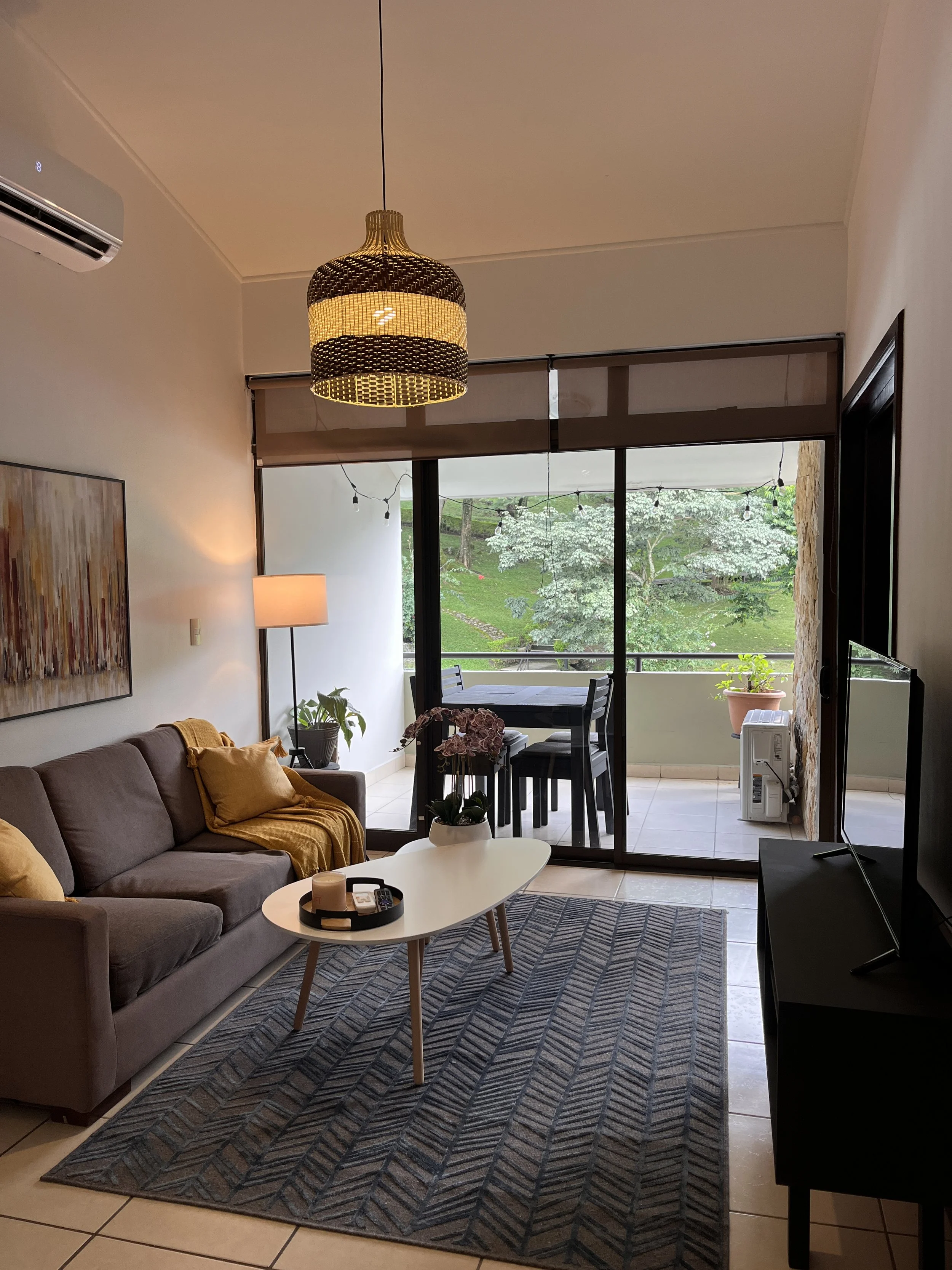 Living room with a gray sofa with yellow cushions, a white oval coffee table with tray, and a patterned rug. A large window opens to a balcony with a table and chairs, overlooking trees. Decor includes a wall painting, a floor lamp, and potted plants. The ceiling has a woven pendant light, and a wall-mounted air conditioner is visible.