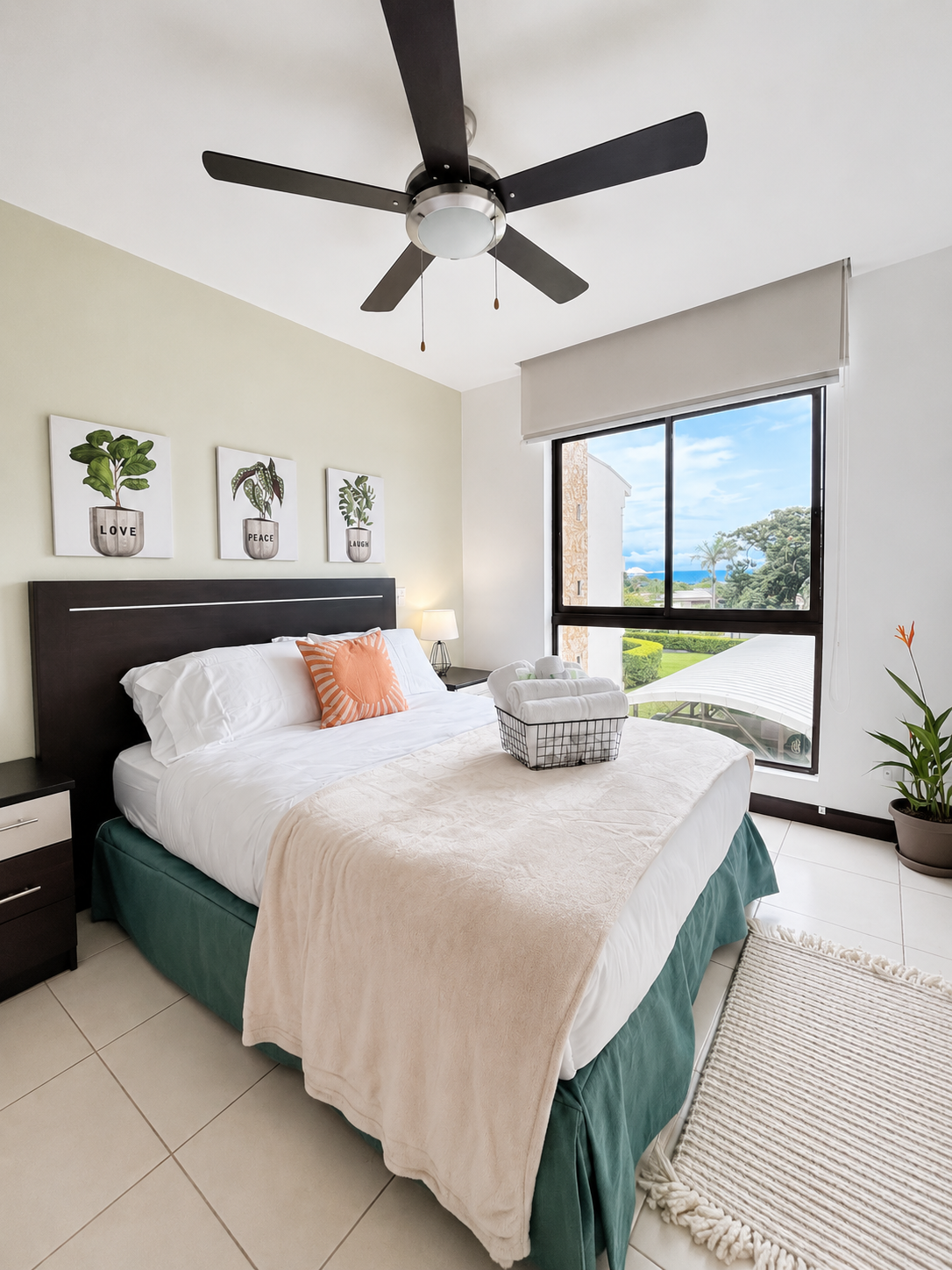 A bedroom with a large bed, dark headboard, white bedding, orange decorative pillow, beige throw blanket, and a basket with rolled towels on the bed. There are three botanical prints on the wall, one with the word 'LOVE', and a window with a view of 