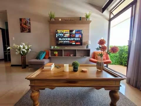 Living room with a wooden coffee table, a flat-screen TV on a wall-mounted unit, a gray sofa with pillows, a vase with white flowers, orange armchair, and large windows with curtains letting in natural light.