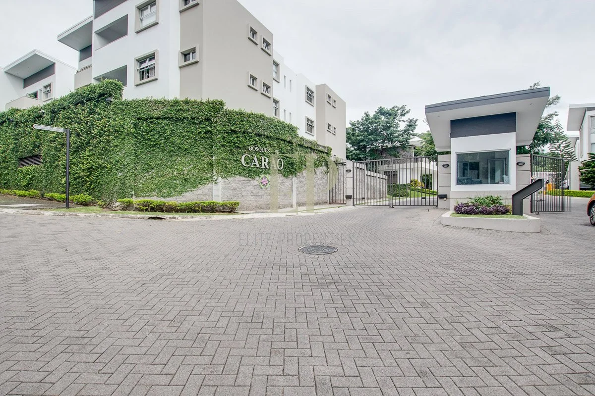 Gated entrance to a modern residential complex with a small security booth and lush greenery in the background.