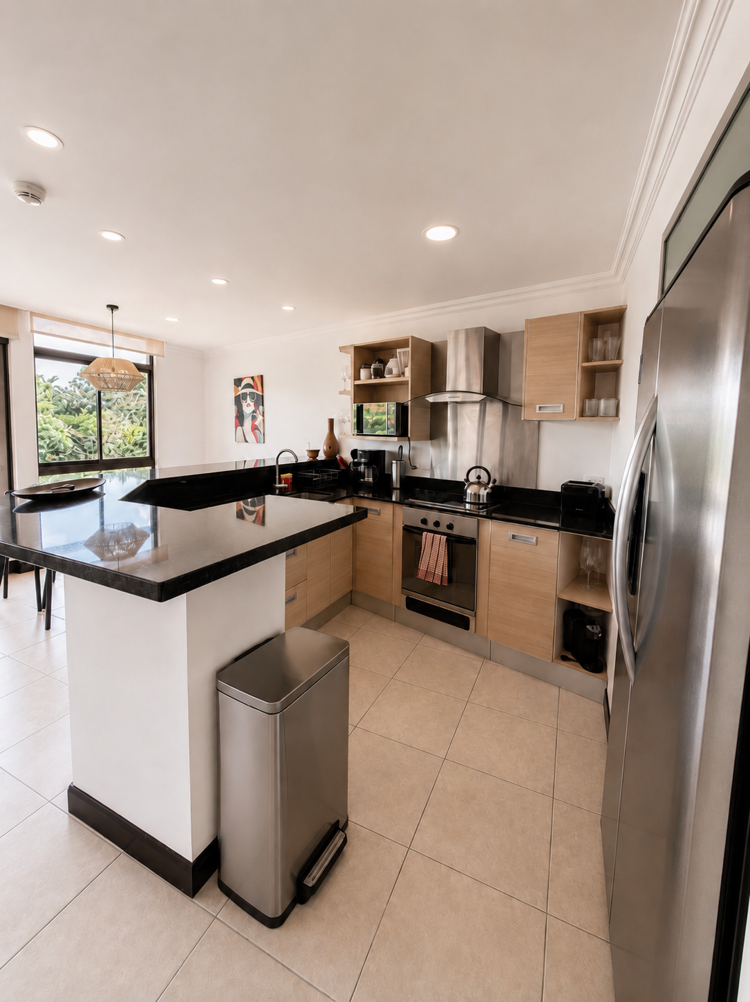 Modern kitchen with black granite countertops, stainless steel appliances, light wood cabinets, and a view of greenery outside through a large window.