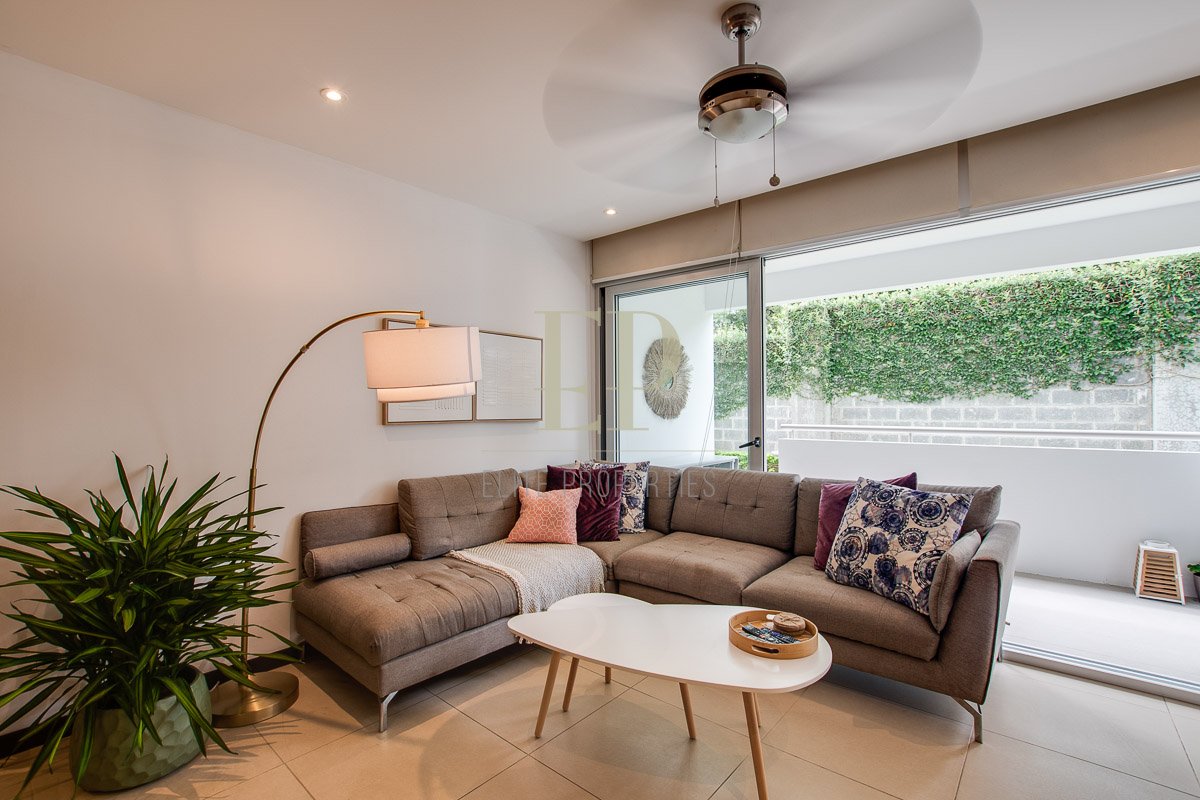 Living room with a beige sectional sofa, colorful throw pillows, a white coffee table, a tall standing lamp, a large window with green foliage outside, and a wall decor piece.