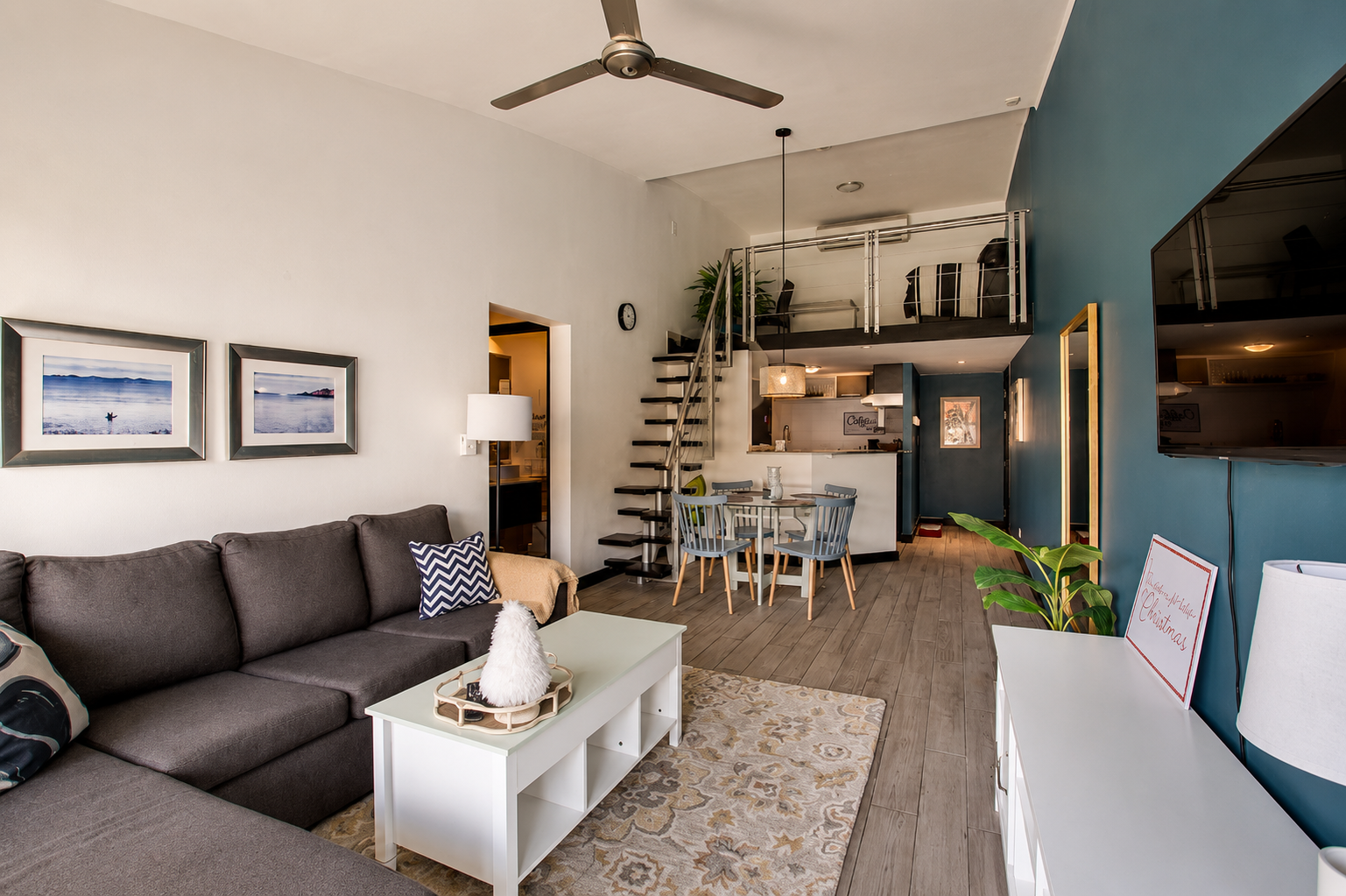 Living room with gray sofa, white coffee table, wall art, indoor potted plant, dining table with four chairs, staircase leading to loft bedroom, television, and Christmas decorations.