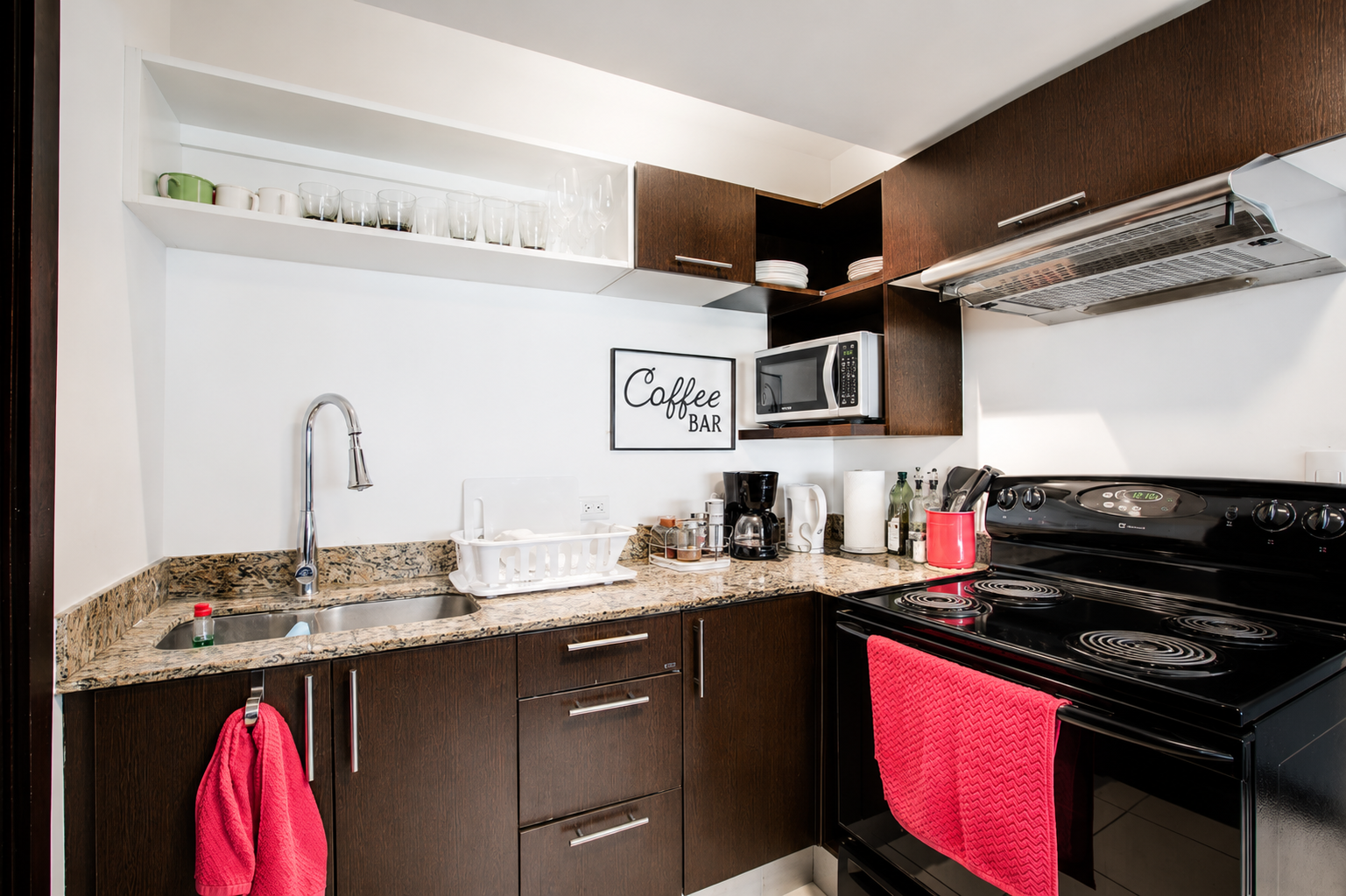 Kitchen with dark wood cabinets, granite countertop, coffee maker, microwave, stove, and red dish towels.