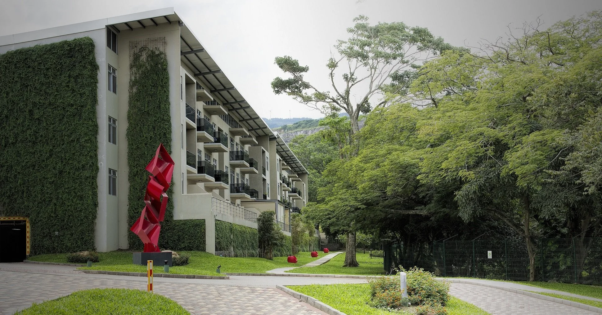 Modern apartment building with balconies, surrounded by lush green trees and a lush landscaped area with abstract red sculpture.