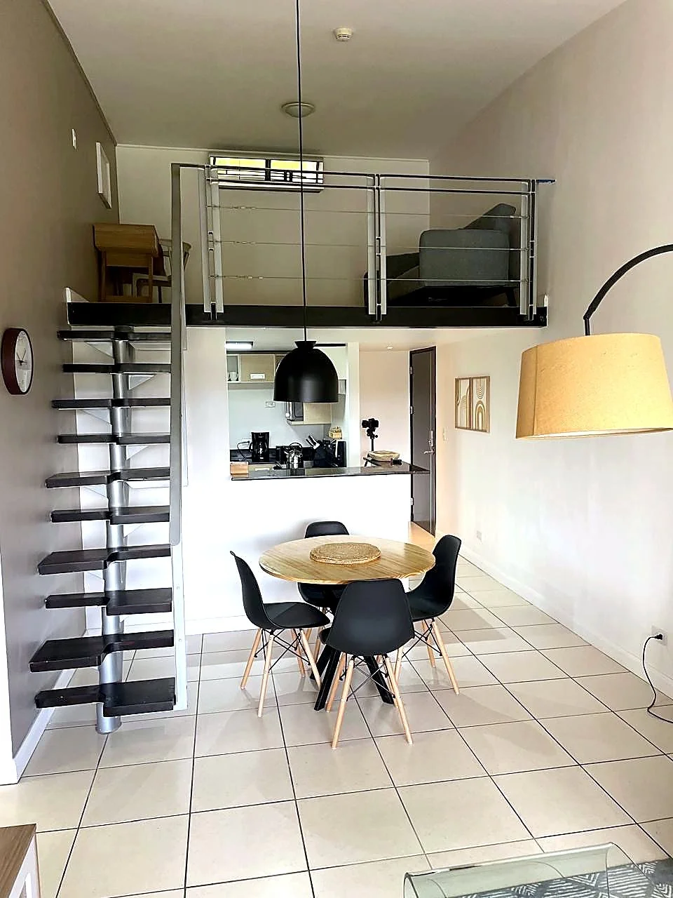 Open concept living space with a dining area, kitchen, and a loft area above. Features include a round wooden table with black chairs, a hanging bamboo shade, a black pendant light, and a spiral staircase leading to the upper loft with sitting and wo