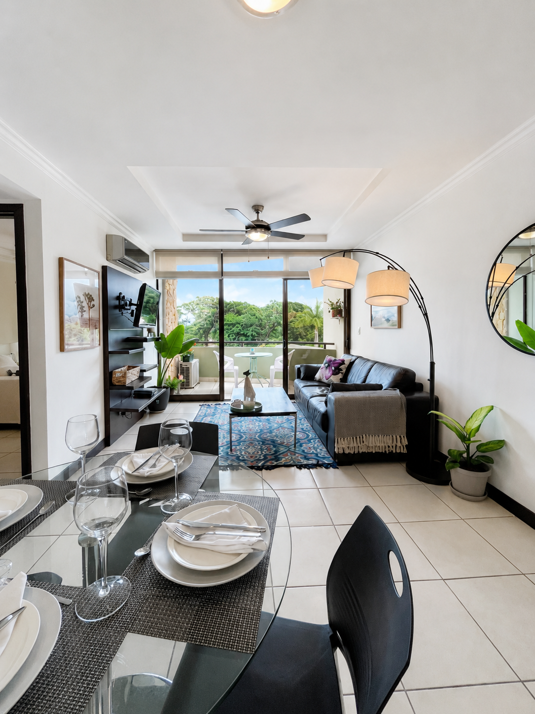 Living room and dining area in an apartment with a balcony, black couch, glass dining table, and large windows with a view of trees.