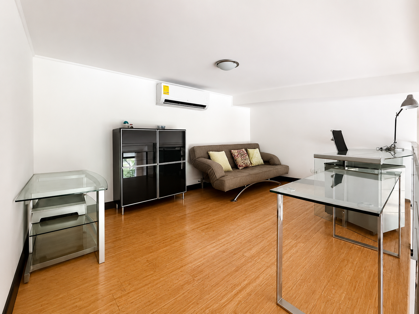 Modern office with wooden floor, white walls, a black cabinet, a beige sofa with cushions, a glass-top desk with a black chair, another glass table with a lamp, and a wall-mounted air conditioner.
