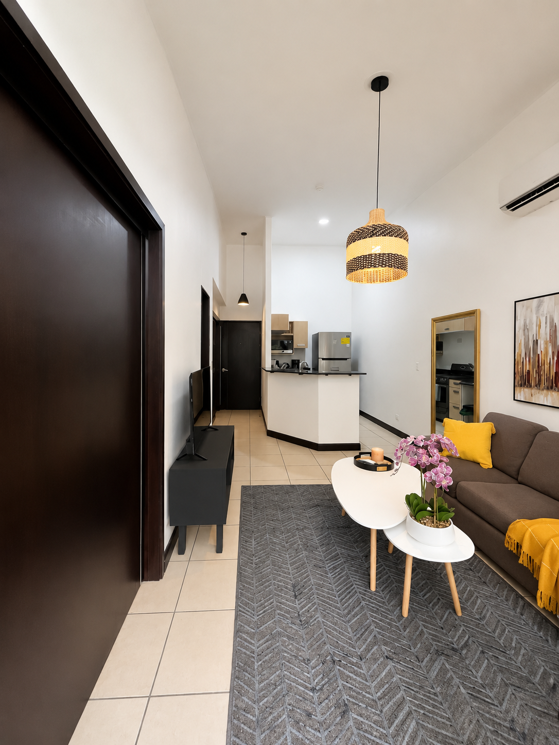 Living room with gray sofa, white oval coffee tables with pink orchids, yellow pillows, a patterned gray rug, wall art, and a kitchen area in the background.