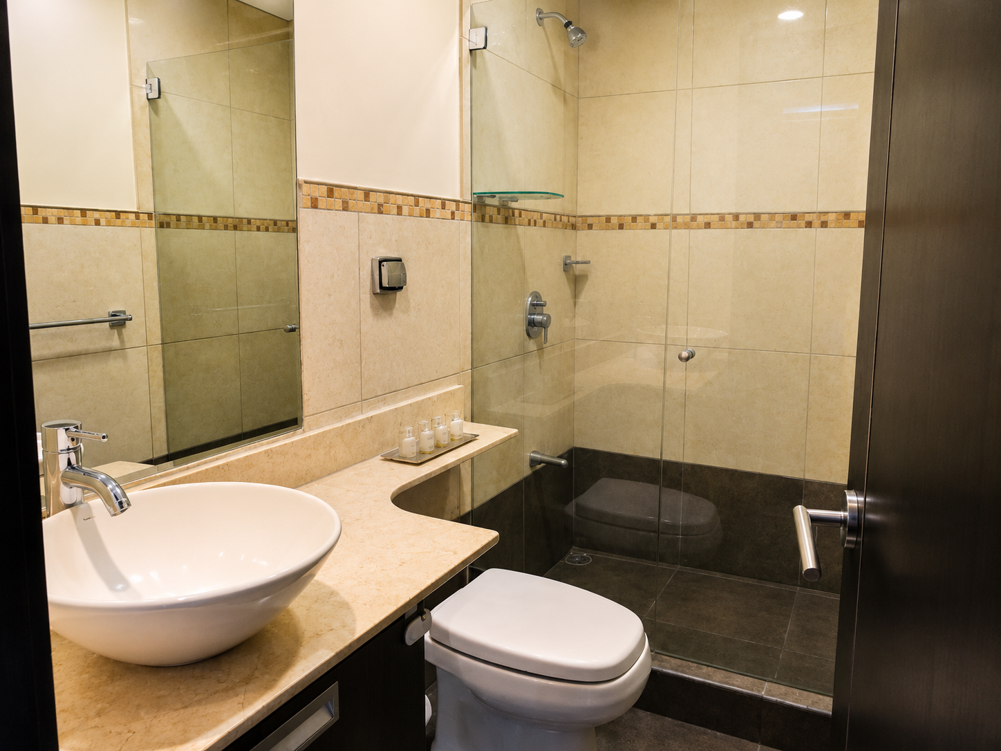 Modern bathroom featuring a glass-enclosed shower with beige and dark tile, a white vessel sink with a chrome faucet on a beige countertop, a mirror, a soap dispenser set, and a toilet next to the shower.