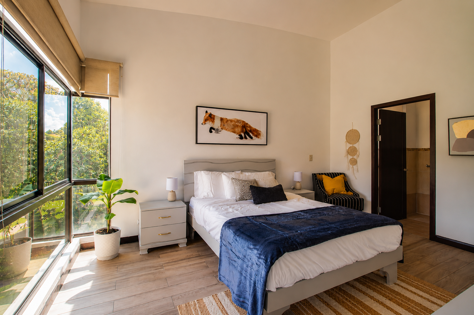 A bedroom with a large bed, white bedding, decorative pillows, and a dark blue throw blanket. There are two white nightstands with lamps, a black and white striped chair with yellow pillows, a potted plant by the window, and a horse-themed wall art a
