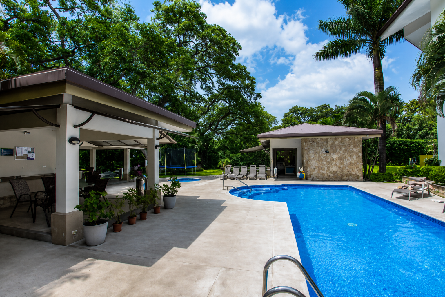 Pool area with lounge chairs, a small pool house with a stone wall, and lush green trees in the background on a sunny day.