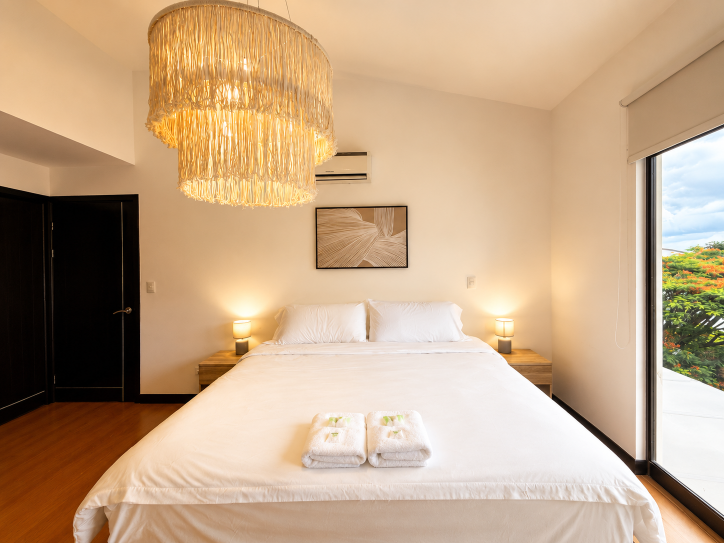 A bedroom with a large white bed, two rolled towels on the bed, two bedside tables with small lamps, a painting above the bed, a large decorative ceiling light, and a sliding glass door that opens to a patio with greenery outside.