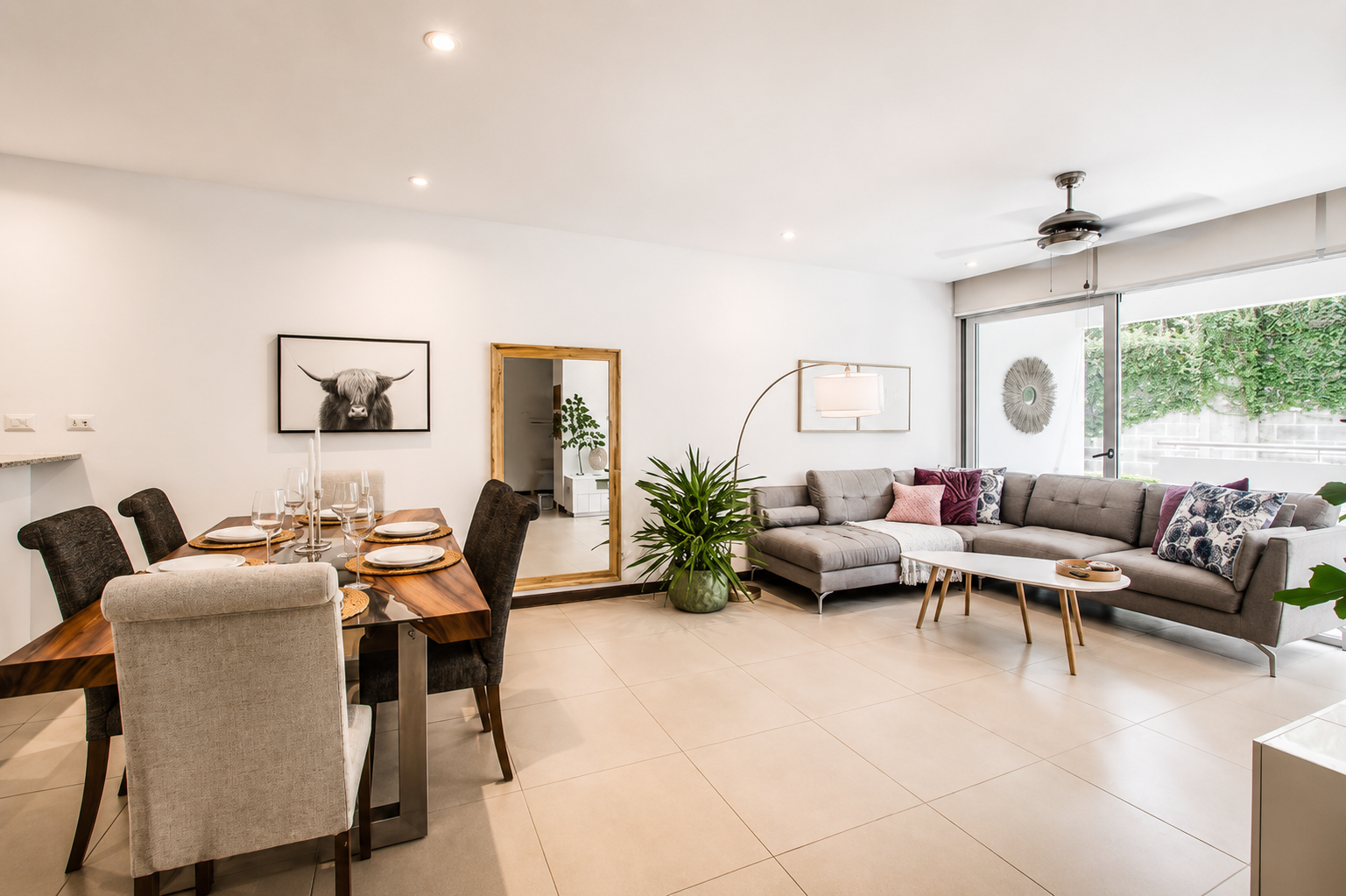 Modern living room and dining area with a large gray sectional sofa, colorful pillows, coffee table, dining table with chairs, a large mirror, wall art, and window view of greenery.