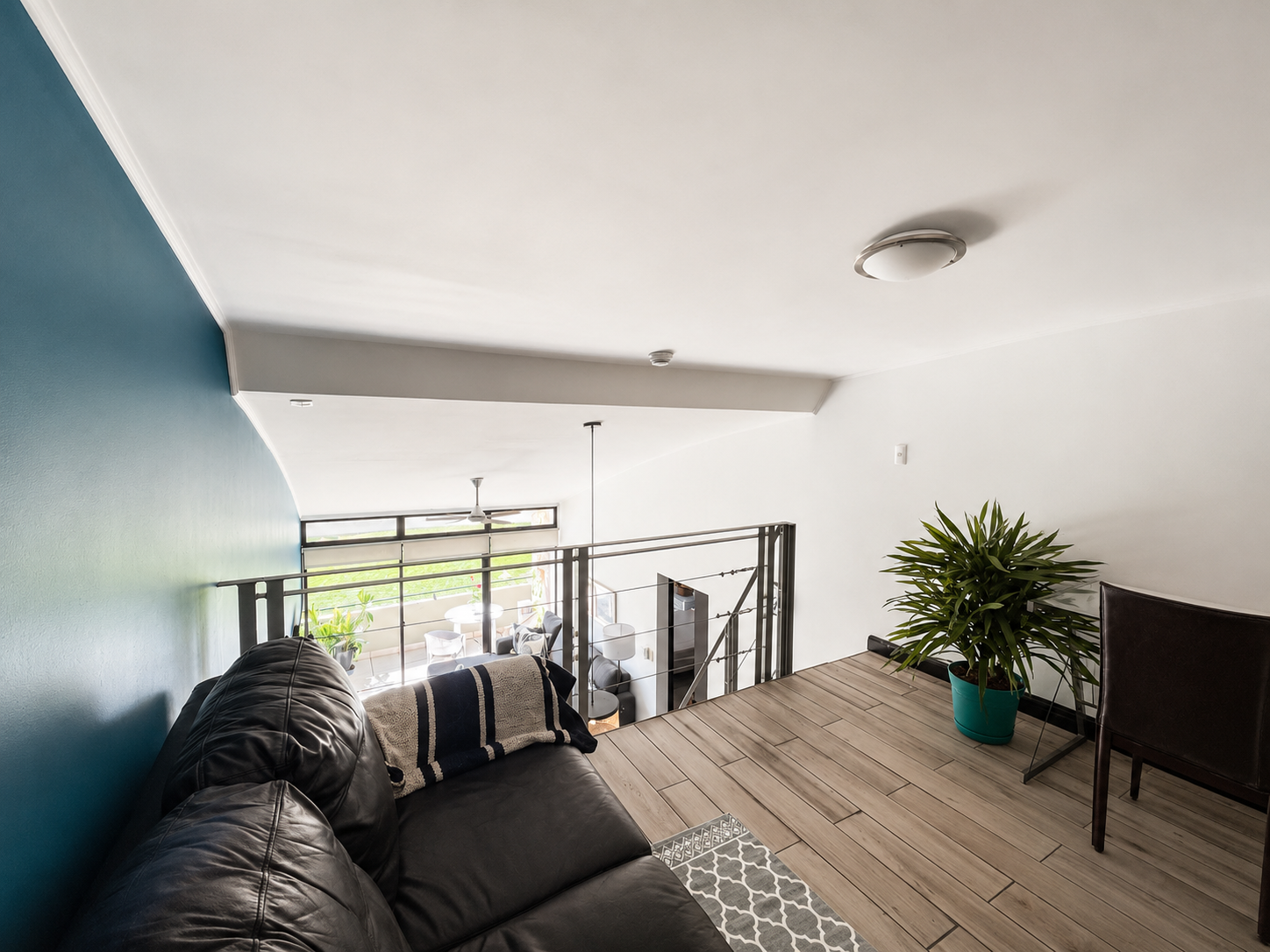 Interior view of a modern living space with a black leather sofa, a blanket, a potted plant, a chair, and a staircase overlooking a lower floor with hanging chairs and large windows letting in natural light.