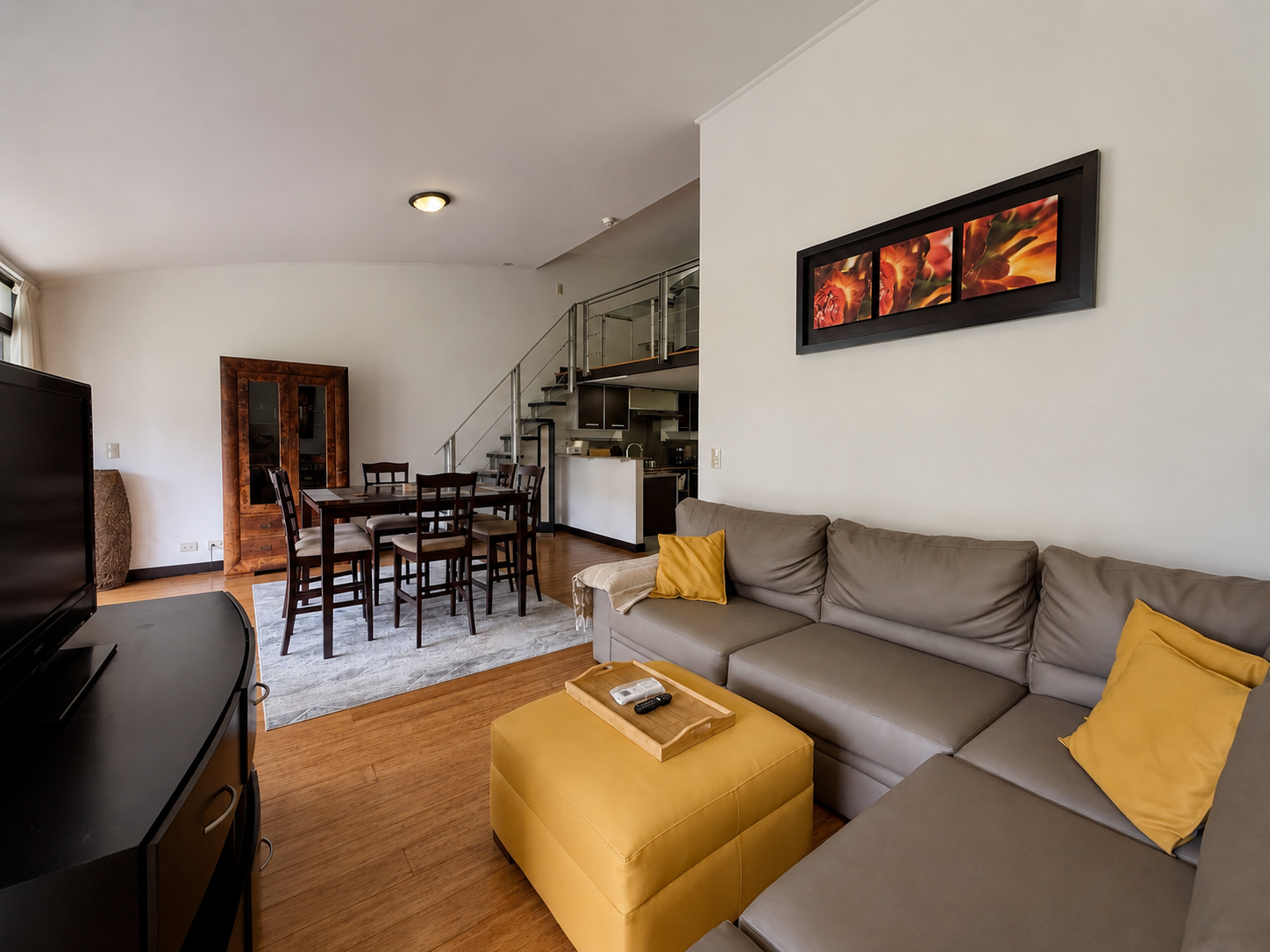 Living room with a beige sectional sofa, yellow pillows, and a yellow ottoman in the foreground, a wooden dining table with six chairs, a staircase leading upstairs, and a kitchen area in the background. A framed floral artwork hangs on the wall.