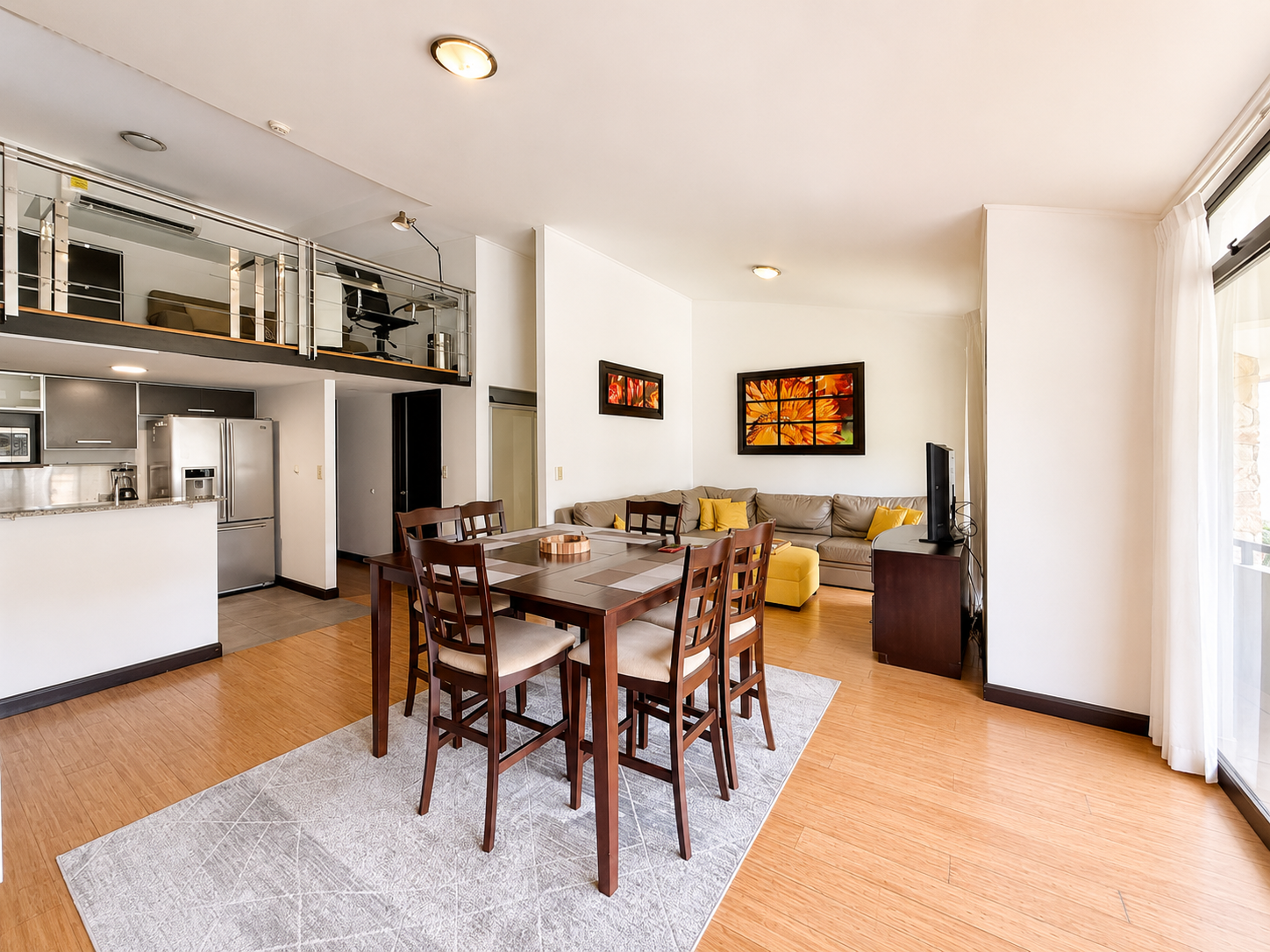 Living room with a dining table, kitchen, and sofa, with artwork on the wall and a TV, overlooking a balcony with sliding glass door.