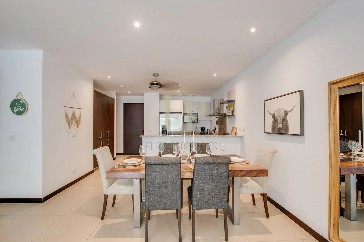 Dining area with a wooden table set for five, six wine glasses, and plates, in a modern home with art and decor on white walls, and a kitchen in the background.