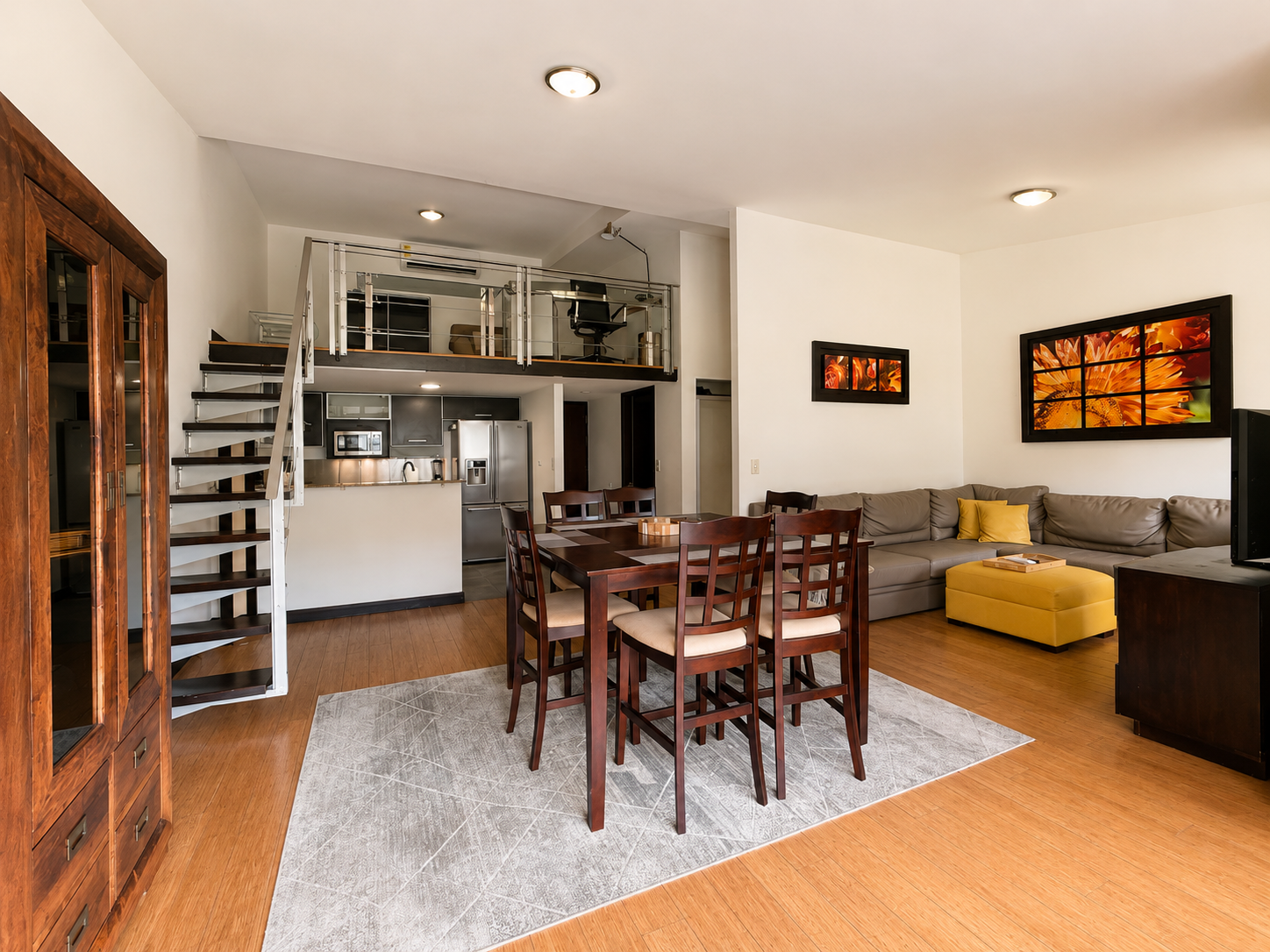 Living room with kitchen, dining area, and loft: wood and beige furniture, artwork, hardwood floors, and a gray sectional couch with yellow accent pillows.