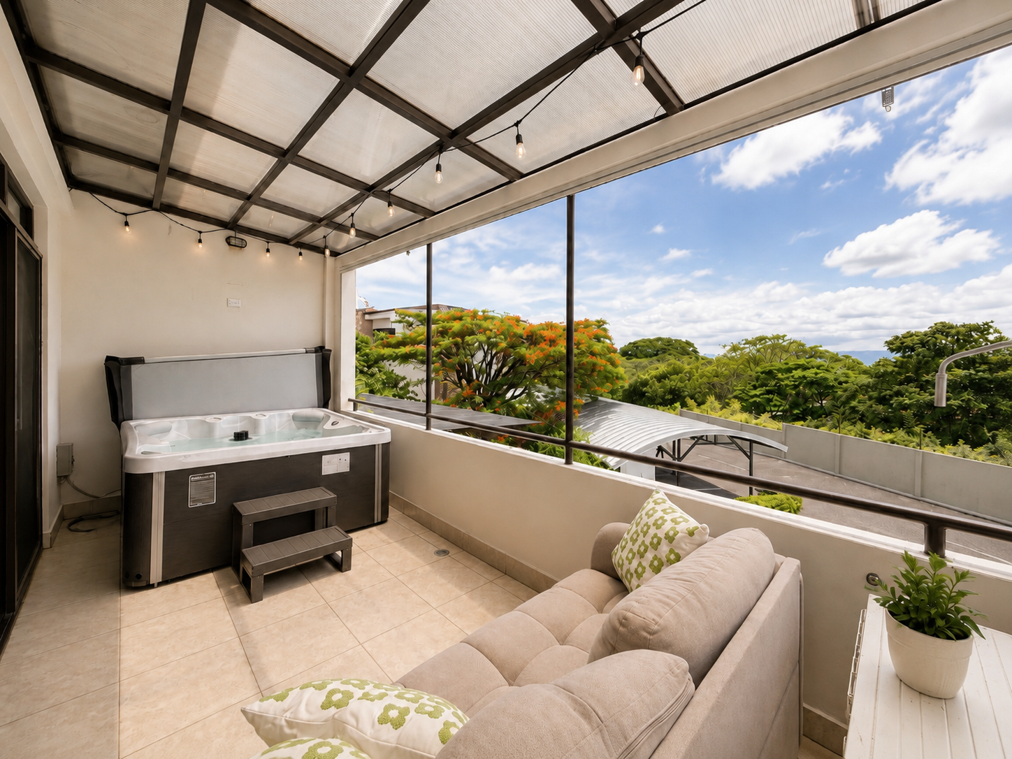 A covered balcony with a hot tub, sofa with green patterned pillows, and a potted plant, overlooking trees and a partly cloudy sky.