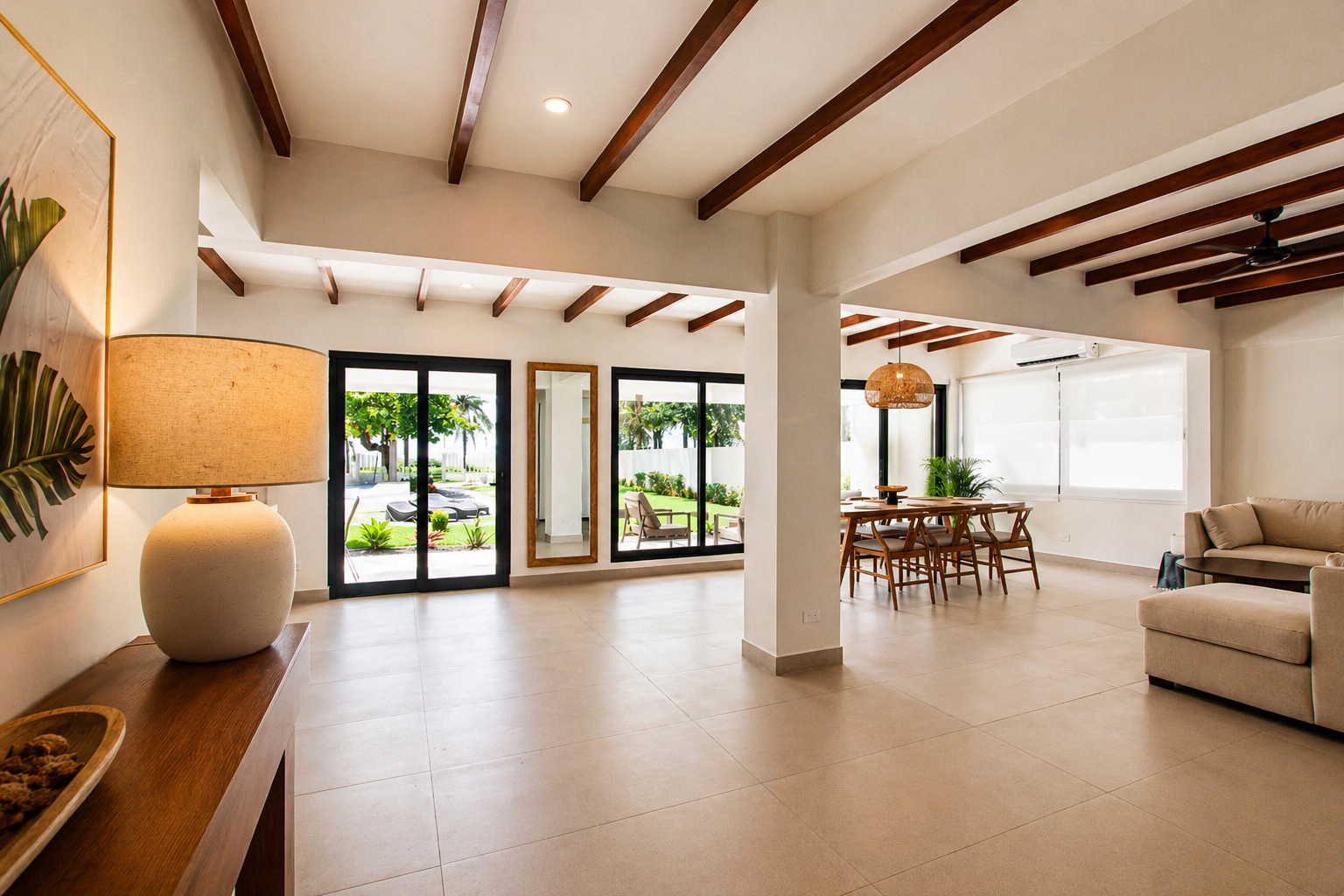 Bright living and dining area with large glass doors, wooden beams on the ceiling, a beige sofa, a dining table with chairs, and tropical outdoor scenery visible through the windows.
