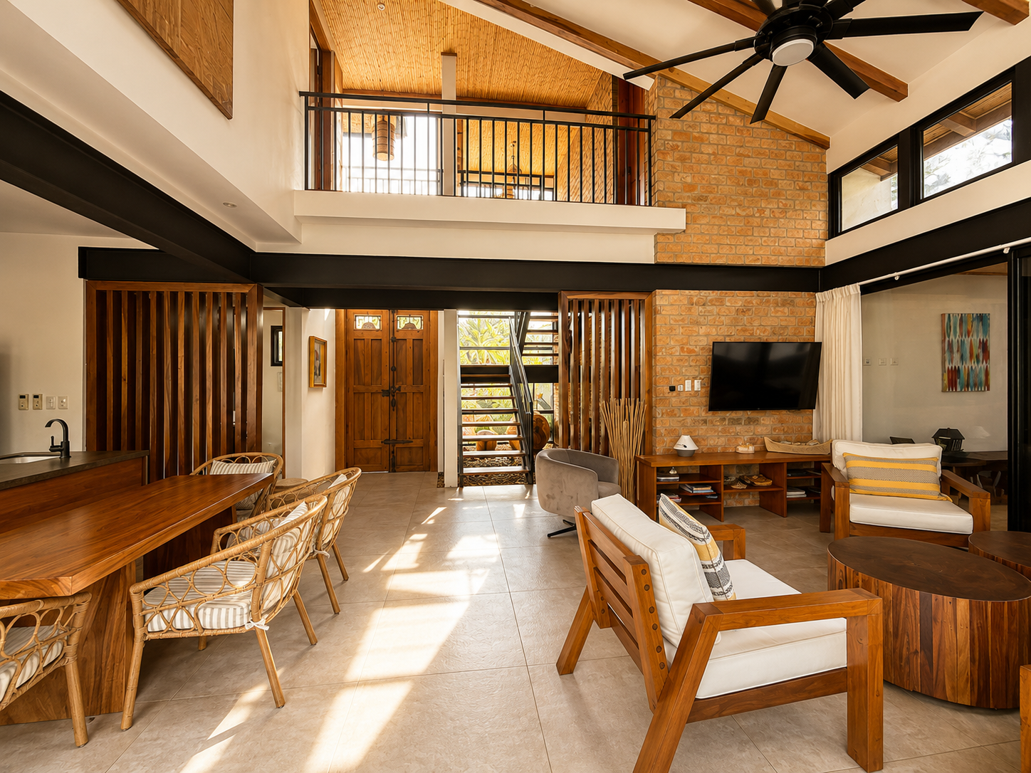 Interior of a modern living room with a high ceiling and a second-floor balcony, featuring wooden accents, a brick wall with a mounted television, a ceiling fan, and large windows letting in natural light.