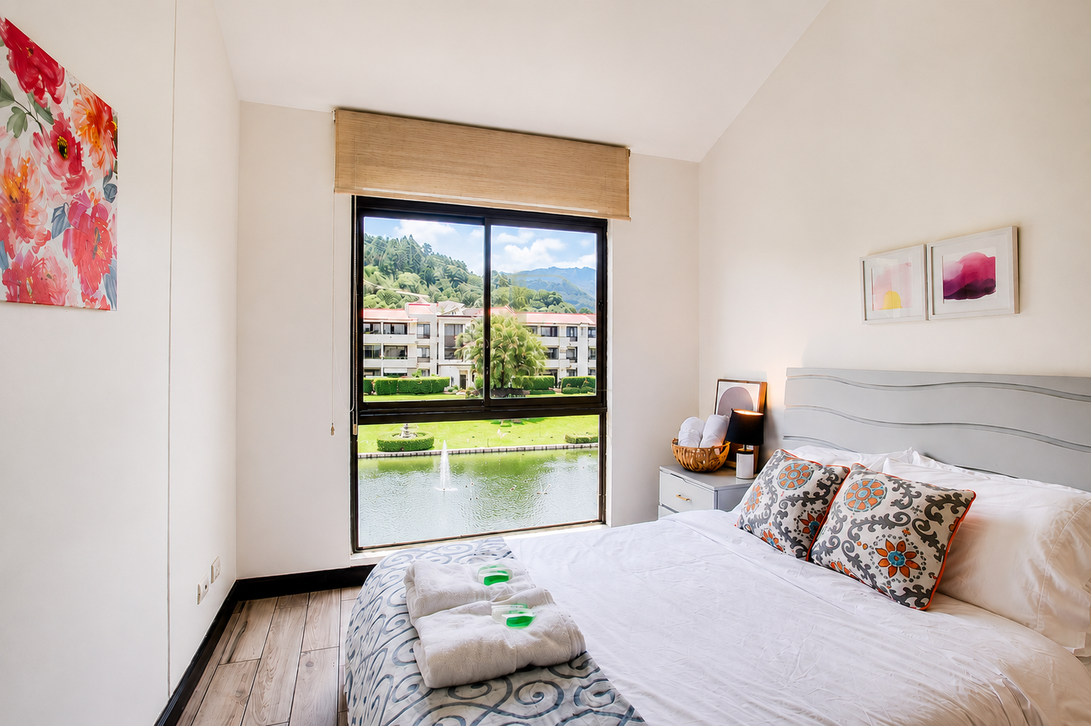 A bedroom with a large window overlooking a pond, fountain, green lawns, apartment complex, trees, and mountains in the background. The bed has two patterned pillows, with towels and toiletries at the foot. Artwork decorates the walls.