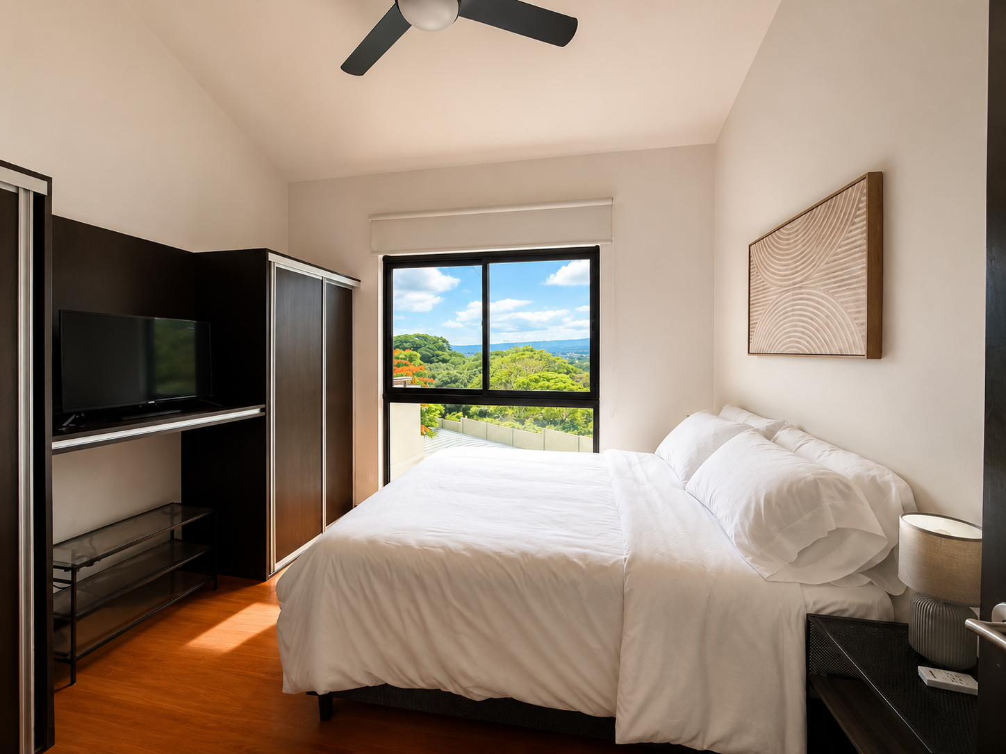 A bedroom with a large bed with white sheets and pillows, a black nightstand with a lamp on it, a black TV on a black wardrobe, a large window showing a scenic view of green trees and hills under a blue sky with clouds, a circular ceiling fan, and a 
