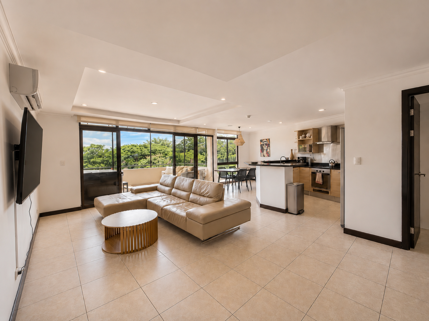 Open living space with beige sectional sofa, small white and wood coffee table, large windows with view of green trees, dining area with black table and chairs, modern kitchen with wooden cabinets, stainless steel appliances, and artwork on the wall.