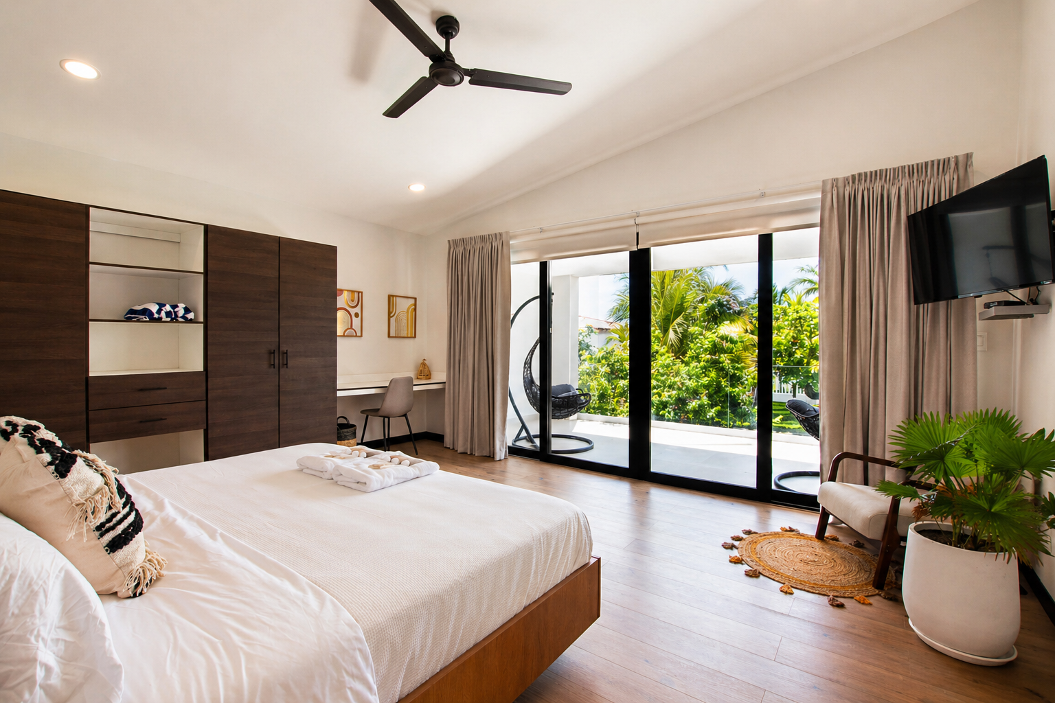 Bright bedroom with sliding glass door opening to a balcony with hanging chair and tropical plants. Contains a large bed, wardrobe, small desk, armchair, and a potted plant.