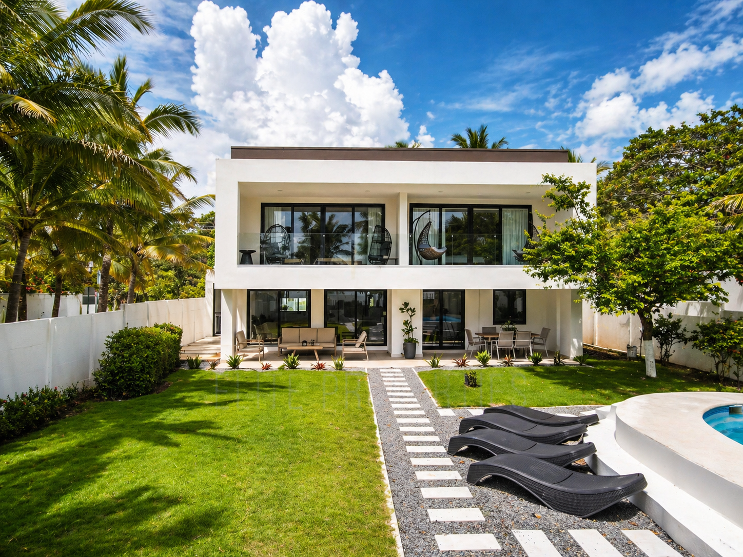 Modern two-story house with white exterior walls, large glass sliding doors, and a balcony with hanging chairs. The front yard features a green lawn, a walking path, and outdoor furniture, with tropical trees and shrubs surrounding the property under