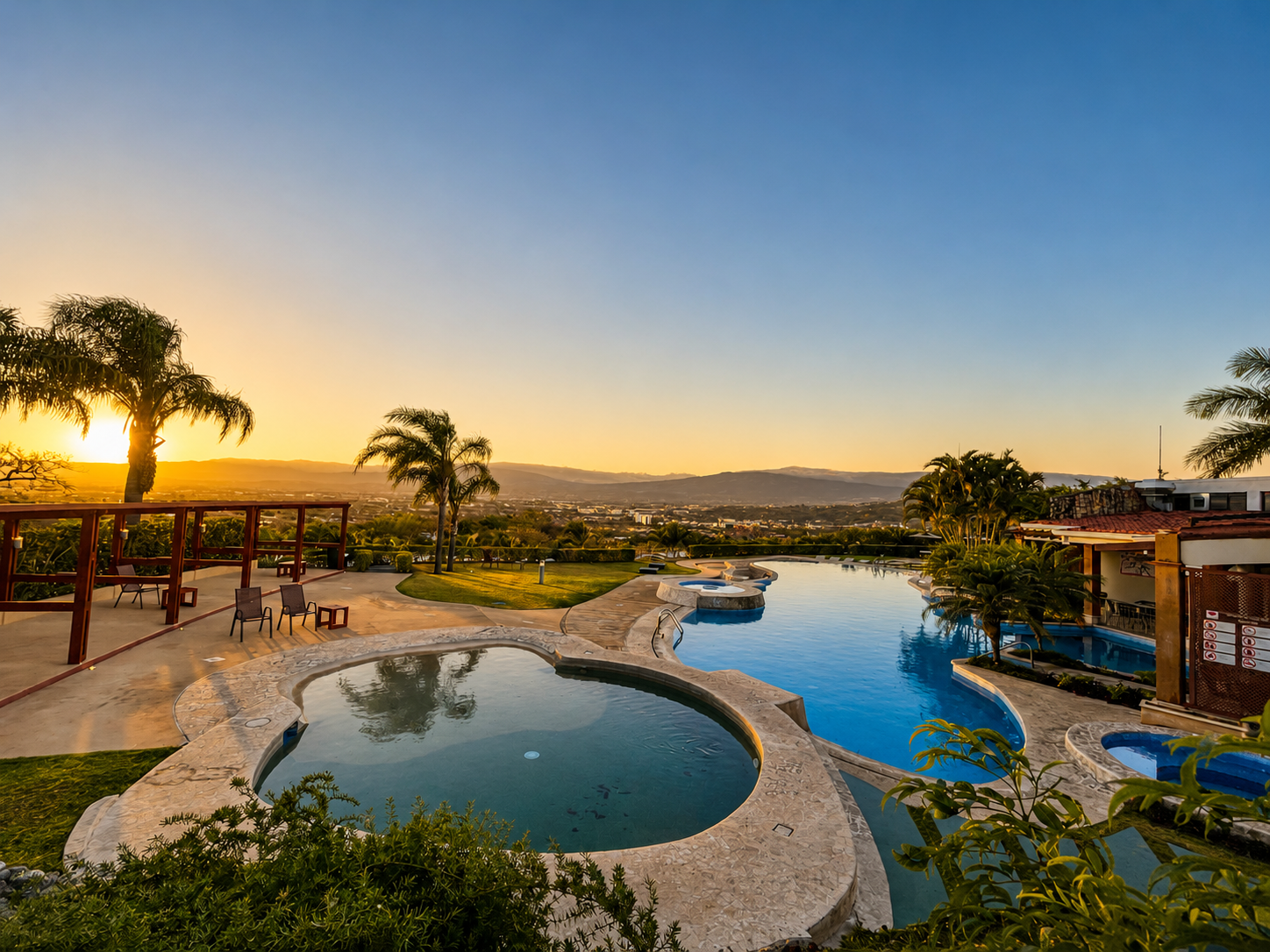 A sunset view over a pool area with palm trees, chairs, and a scenic landscape in the background.