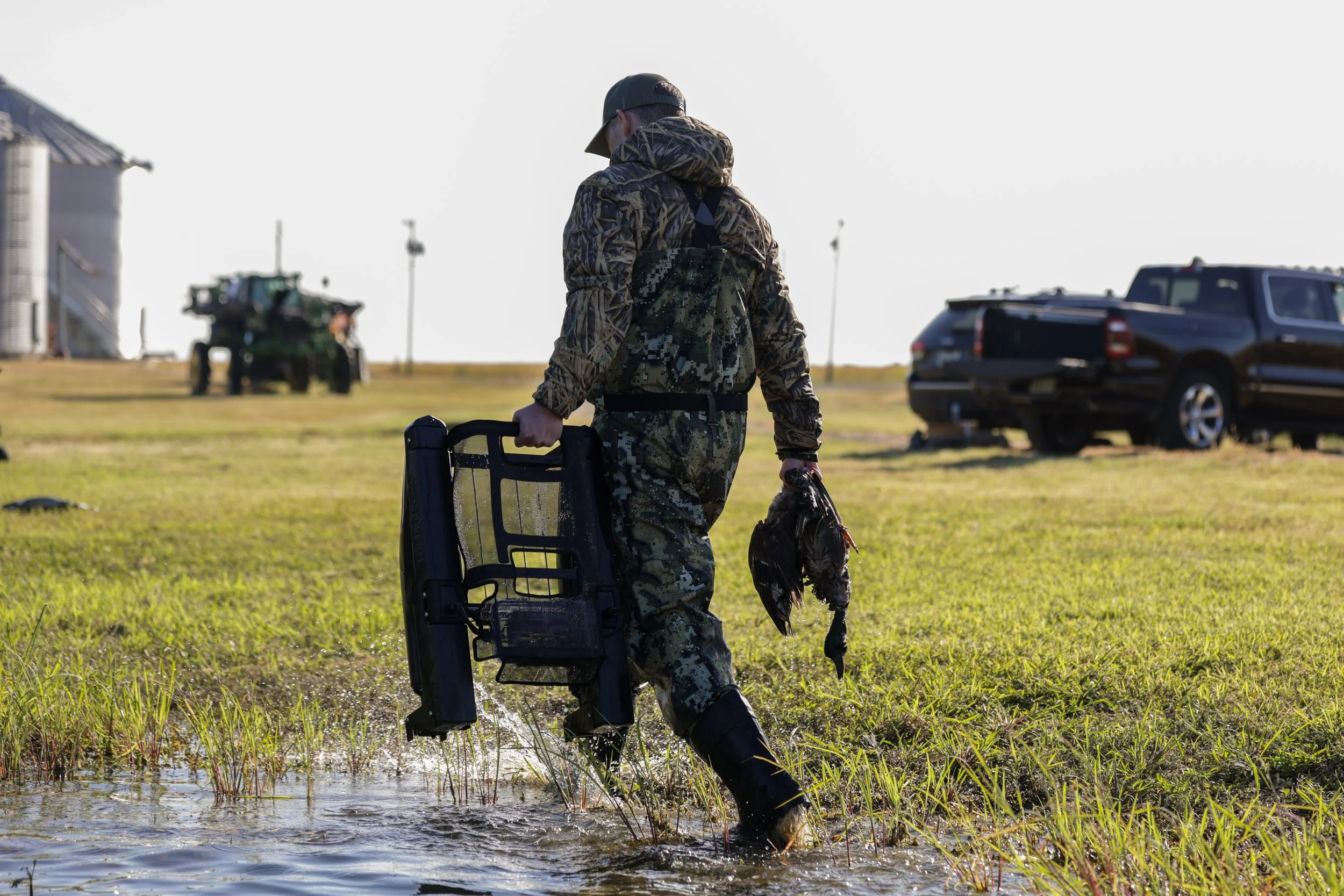 A person dressed in camouflage hunting gear and a cap walking through a marshy area, carrying a plastic bag with a dead duck, with two pickup trucks and an industrial building in the background.