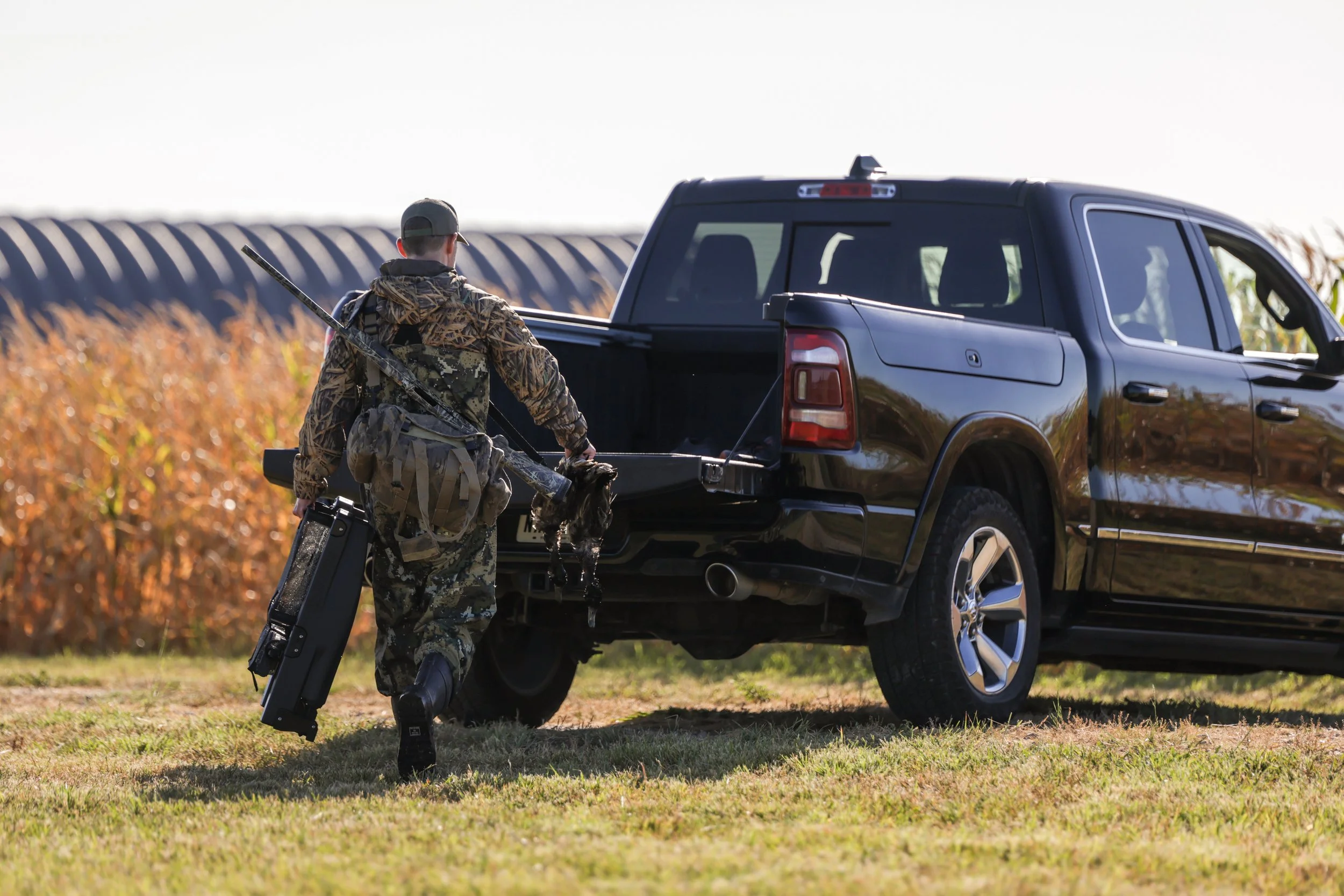 A person in camouflage hunting clothing walking towards a black pickup truck in a field with autumn-colored bushes in the background. The person is carrying a rifle and a Fetch Waterfowl Retrieval System.