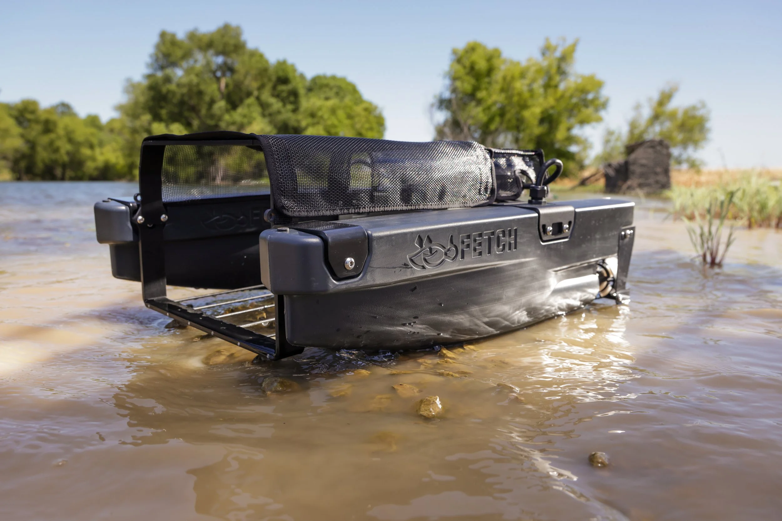 A picture of the Fetch waterfowl retrieval system in shallow water atop some rocks, surrounded by trees and a clear blue sky in the background.